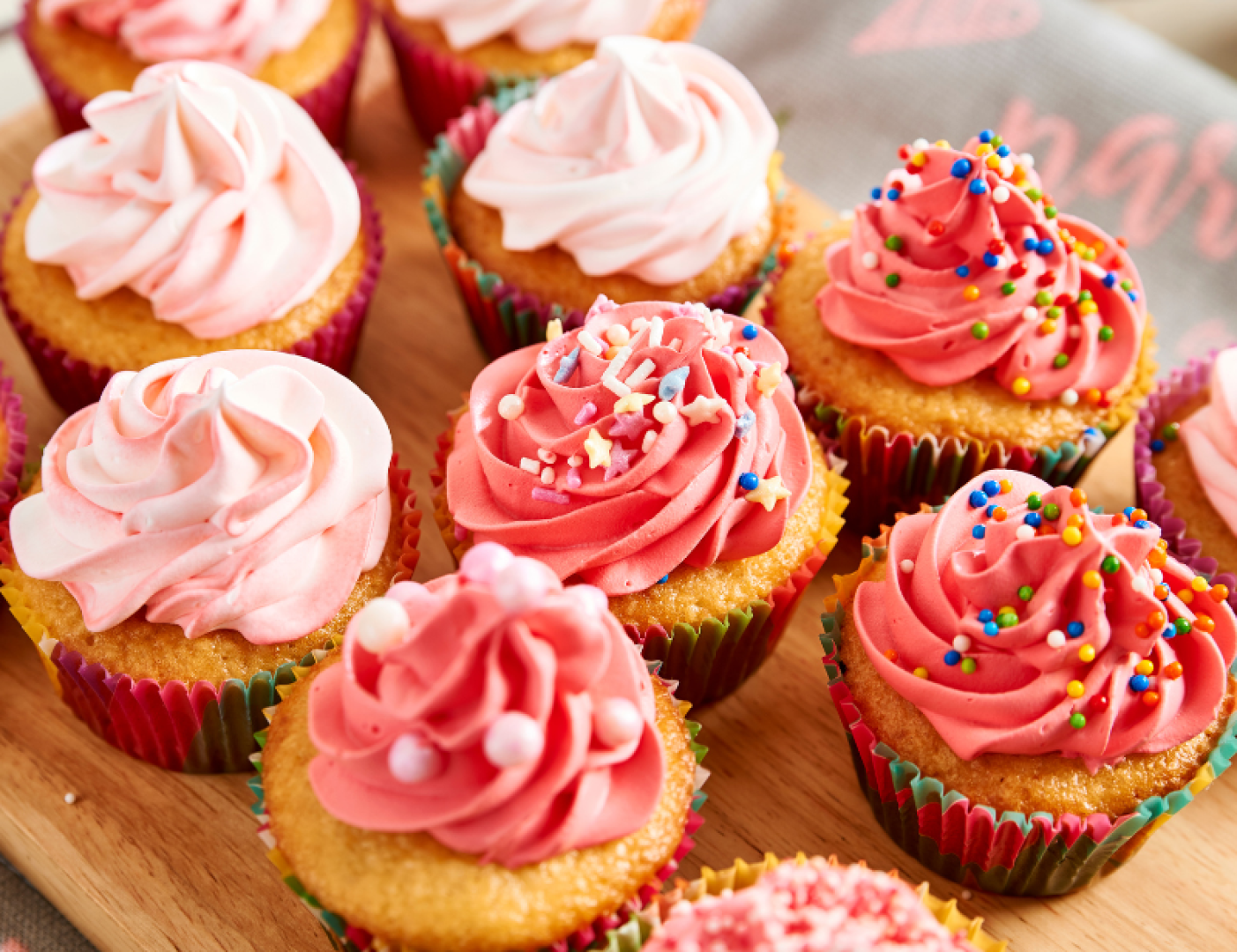 Eine Auswahl an Cupcakes mit rosa und rotem Zuckerguss, einige davon mit bunten Streuseln bestreut, dicht aneinandergereiht auf einer Holzfläche.
