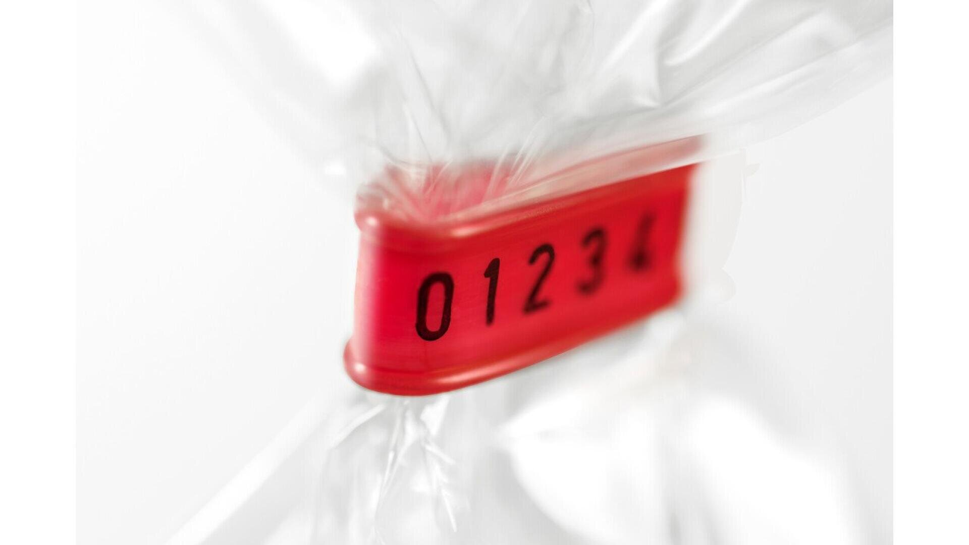 Nahaufnahme eines roten Sicherheitssiegels aus Kunststoff mit den aufgedruckten Zahlen 01234, das um eine durchsichtige Plastiktüte befestigt ist. Der Hintergrund ist weiß und unscharf.