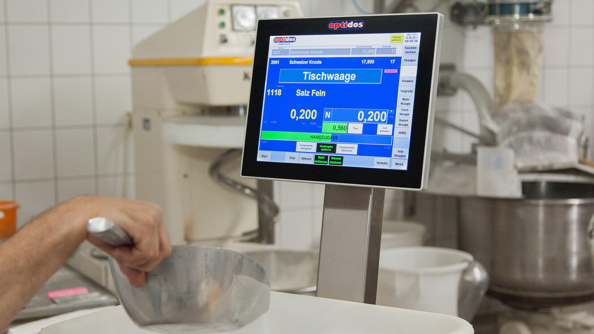 Eine Person schöpft feines Salz in einen Behälter auf einer digitalen Waage mit einem Display, das die Gewichtsmessungen in einer gekachelten Industrieküche anzeigt.