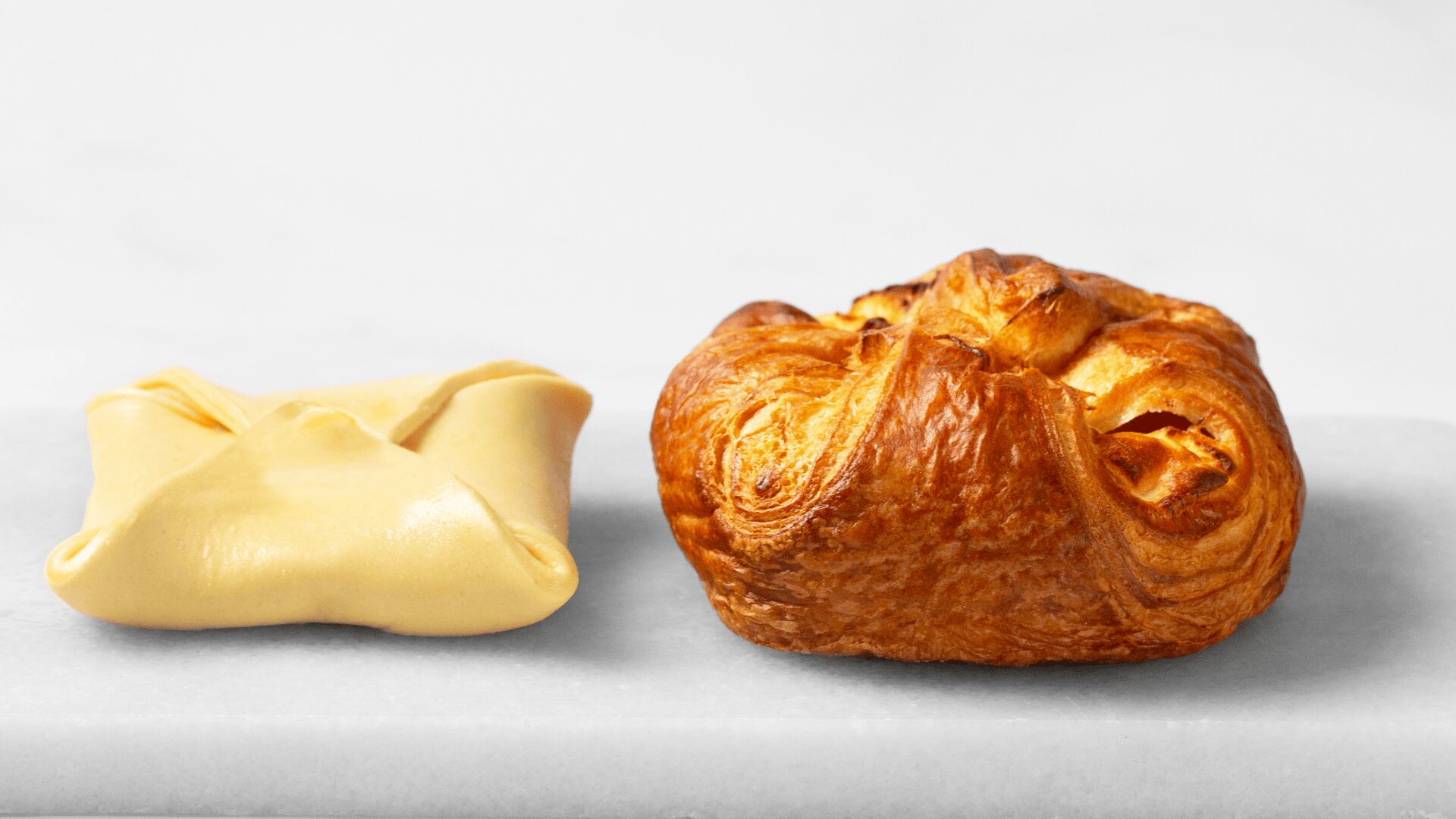 Zwei Gebäckstücke auf einer weißen Marmorplatte: das eine ist ungebacken und hat einen glatten, gefalteten Teig, das andere ist goldbraun gebacken und hat eine runde, aufgeblasene Form.