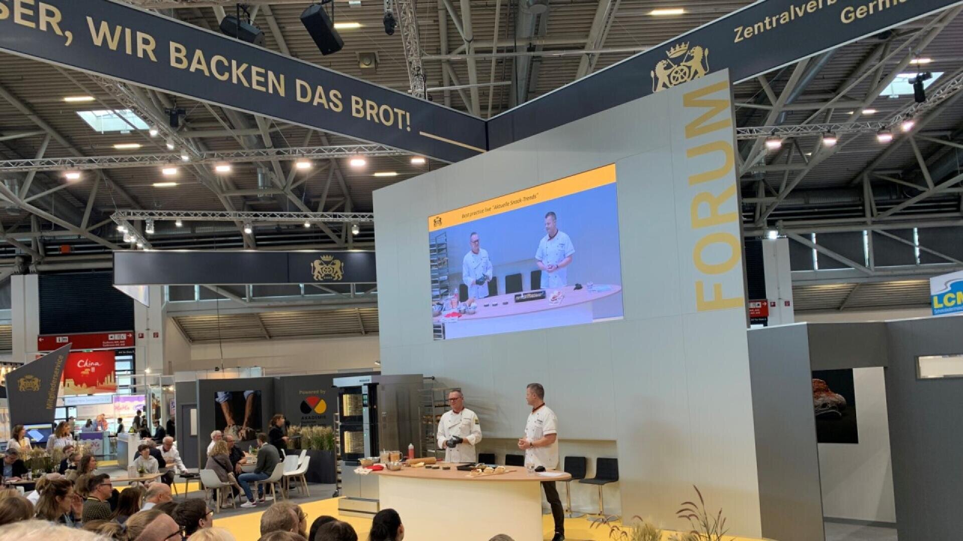 Two people in white uniforms give a baking demonstration on stage at a forum, with an audience watching and a large screen behind them displaying their actions. Overhead, a banner reads, “WIR BACKEN DAS BROT!” in German.
