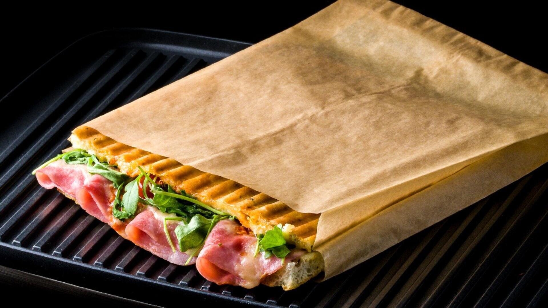Ein gegrilltes Panini-Sandwich mit Schinken, Käse und Blattgemüse wird teilweise in braunes Pergamentpapier eingewickelt und auf einen schwarzen Grill gelegt.
