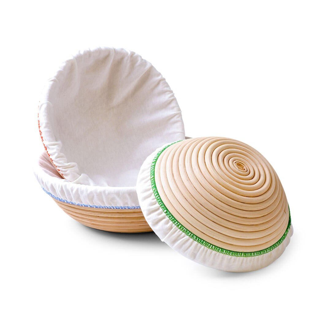 Zwei runde, hölzerne, mit weißem Stoff ausgekleidete Gärkörbe, einer offen und einer mit schräg aufliegendem Deckel. Die Körbe haben spiralförmige Rillen und dekorative Nähte an den Rändern des Stoffes.
