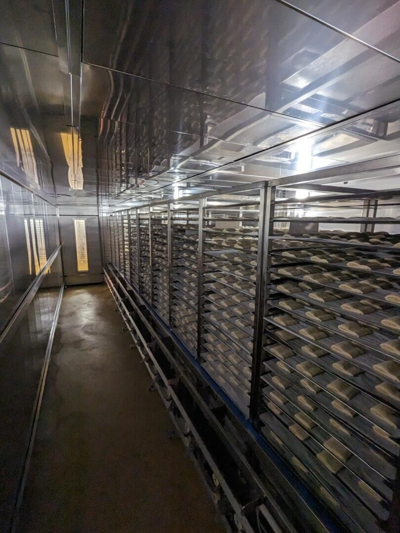 Rows of metal racks filled with trays of dough or bread sit in a long, narrow, industrial kitchen or bakery space with reflective metallic walls and ceiling.