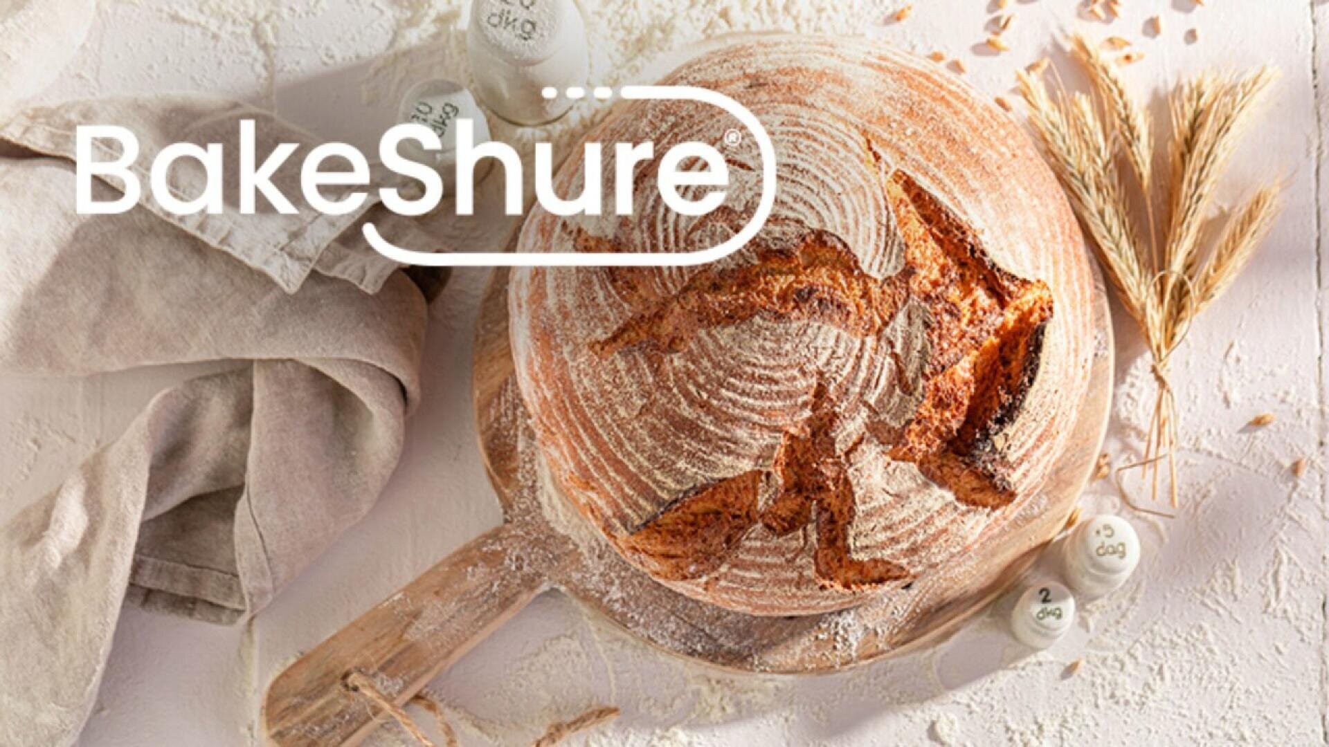 Ein runder Laib rustikales Brot auf einem Holzbrett, mit Mehl bestreut, daneben Weizenhalme, ein beiges Tuch und Messlöffel. Das Wort „BakeShure“ ist in fettem weißen Text darübergelegt.