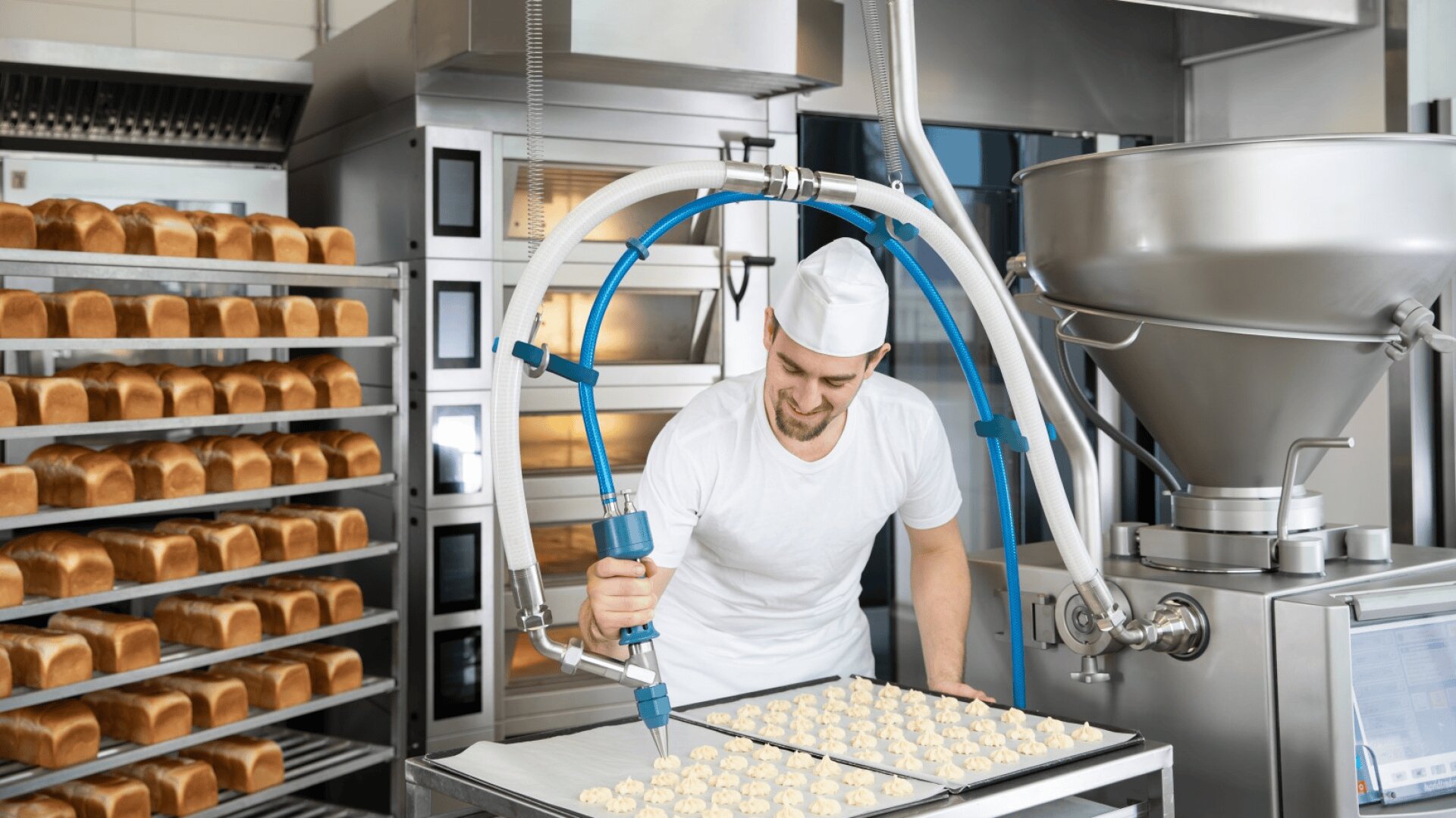 Ein Bäcker in weißer Uniform und mit Mütze benutzt eine Maschine, um Teig auf ein Tablett in einer gewerblichen Bäckerei zu geben, wobei im Hintergrund Regale mit Brotlaiben zu sehen sind.