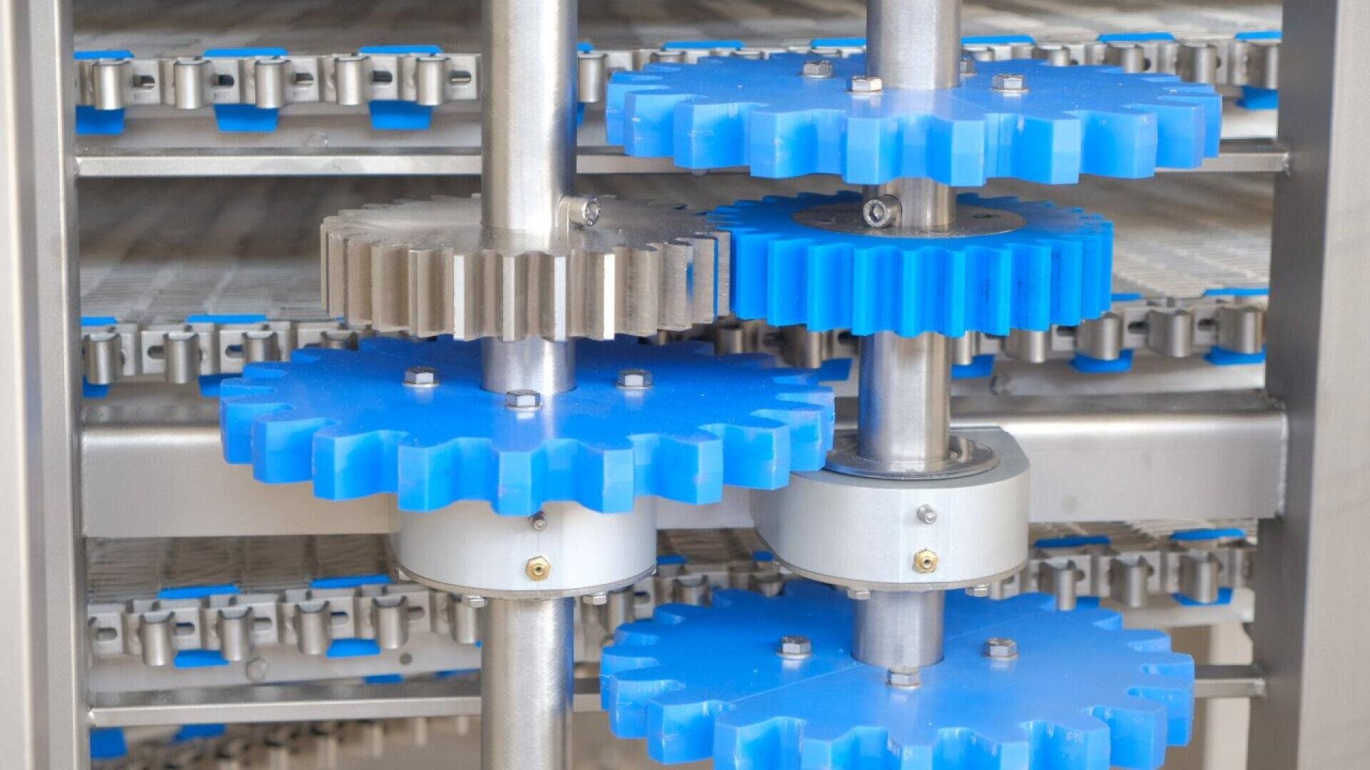 Nahaufnahme mehrerer blauer und silberner Kunststoffzahnräder, die auf Metallstangen in einer Industriemaschine montiert sind, wobei im Hintergrund Förderbänder und weitere Maschinenteile zu sehen sind.