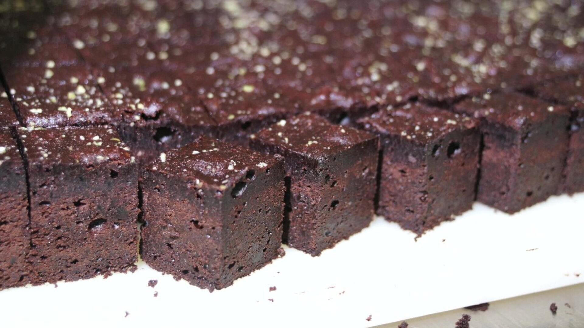 Rows of neatly cut, square chocolate brownies are arranged close together on a white surface, with a slight dusting of sugar or powder on top. The brownies appear moist and rich in color.
