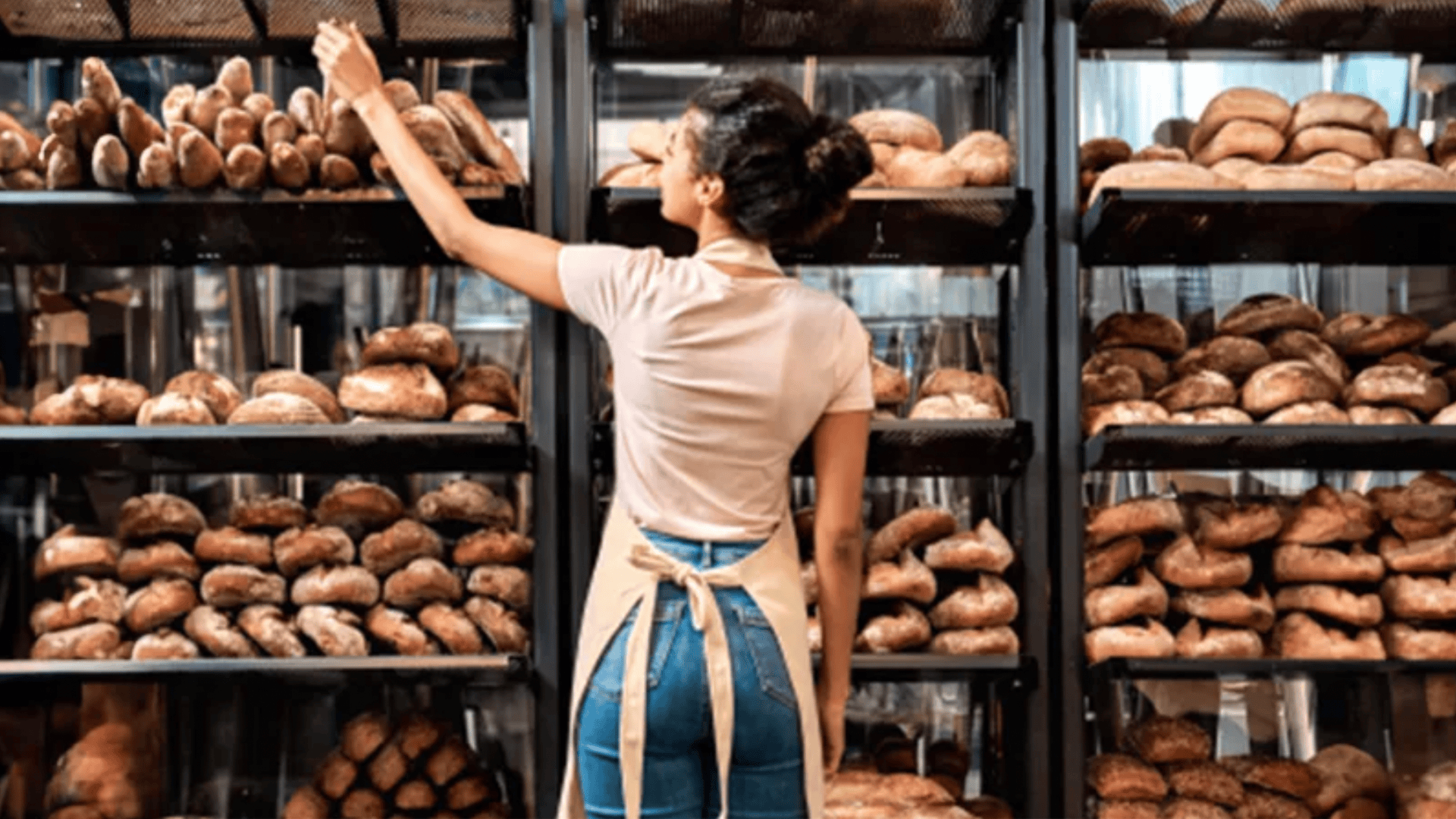 Eine Frau mit Schürze ordnet Brote in Regalen in einer Bäckerei an, wobei sie der Kamera den Rücken zuwendet und von Regalen mit verschiedenen Brotsorten umgeben ist.