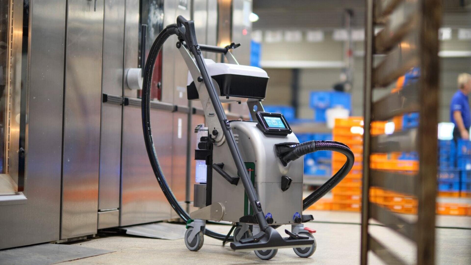 A compact, wheeled cleaning robot with attached hoses and a touchscreen stands in an industrial setting near metallic equipment and shelving with orange crates in the background.