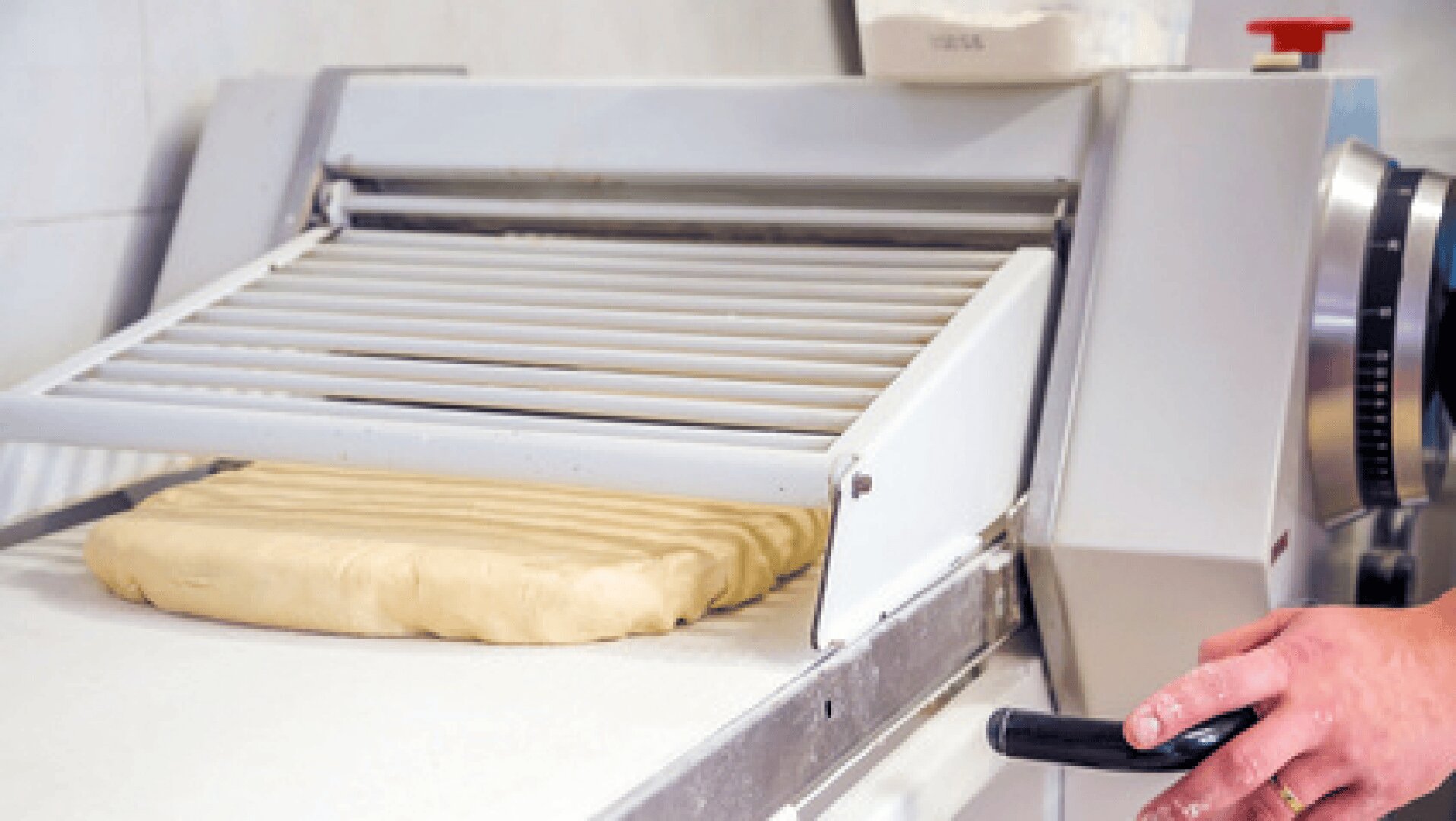 Eine Person bedient eine Teigausrollmaschine, die eine Teigplatte auf einer weißen Fläche in einer Großküche oder Bäckerei ausrollt.