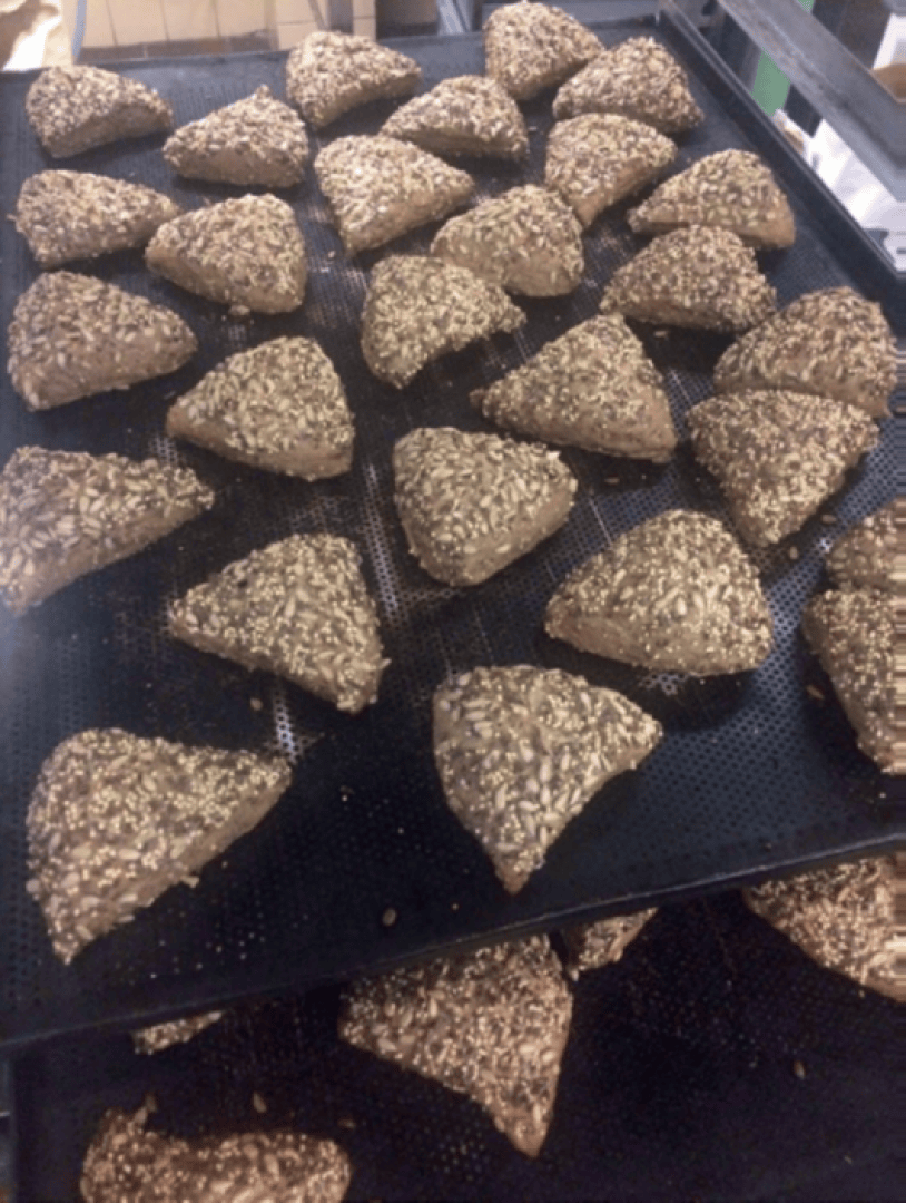 Several triangular, brown bread rolls topped with seeds are arranged in rows on a black baking tray, ready for baking.