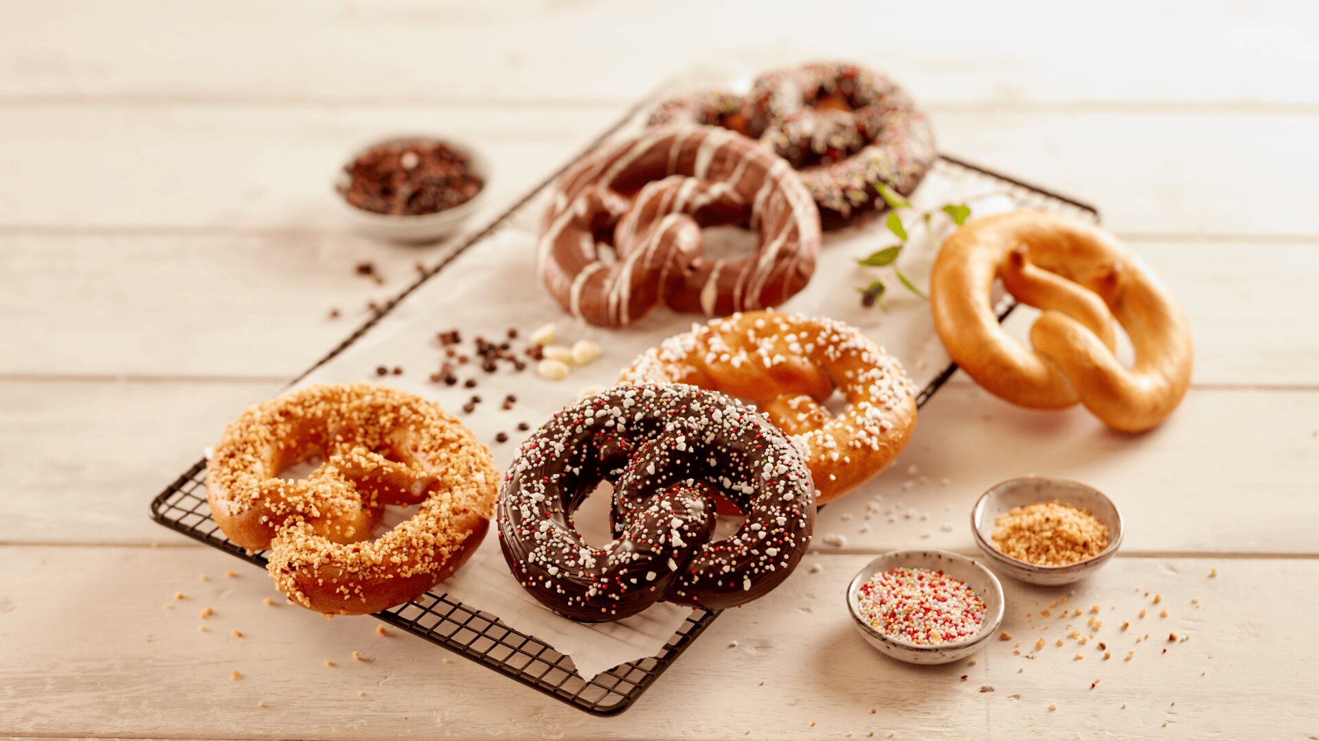 Assorted pretzels, some chocolate-coated and sprinkled with toppings, sit on a cooling rack and white wooden table, surrounded by bowls of colorful sprinkles and crushed nuts.