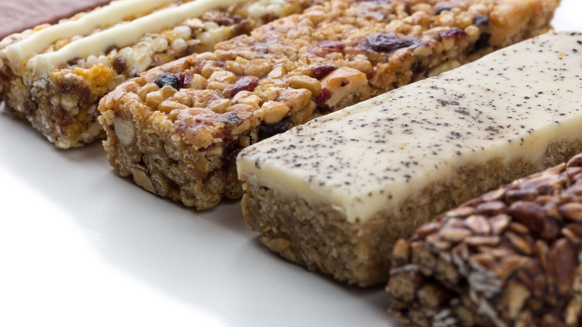 Eine Nahaufnahme mehrerer verschiedener Müsli- und Proteinriegel, jeweils mit unterschiedlichen Belägen und Texturen, nebeneinander auf einer weißen Oberfläche angeordnet.