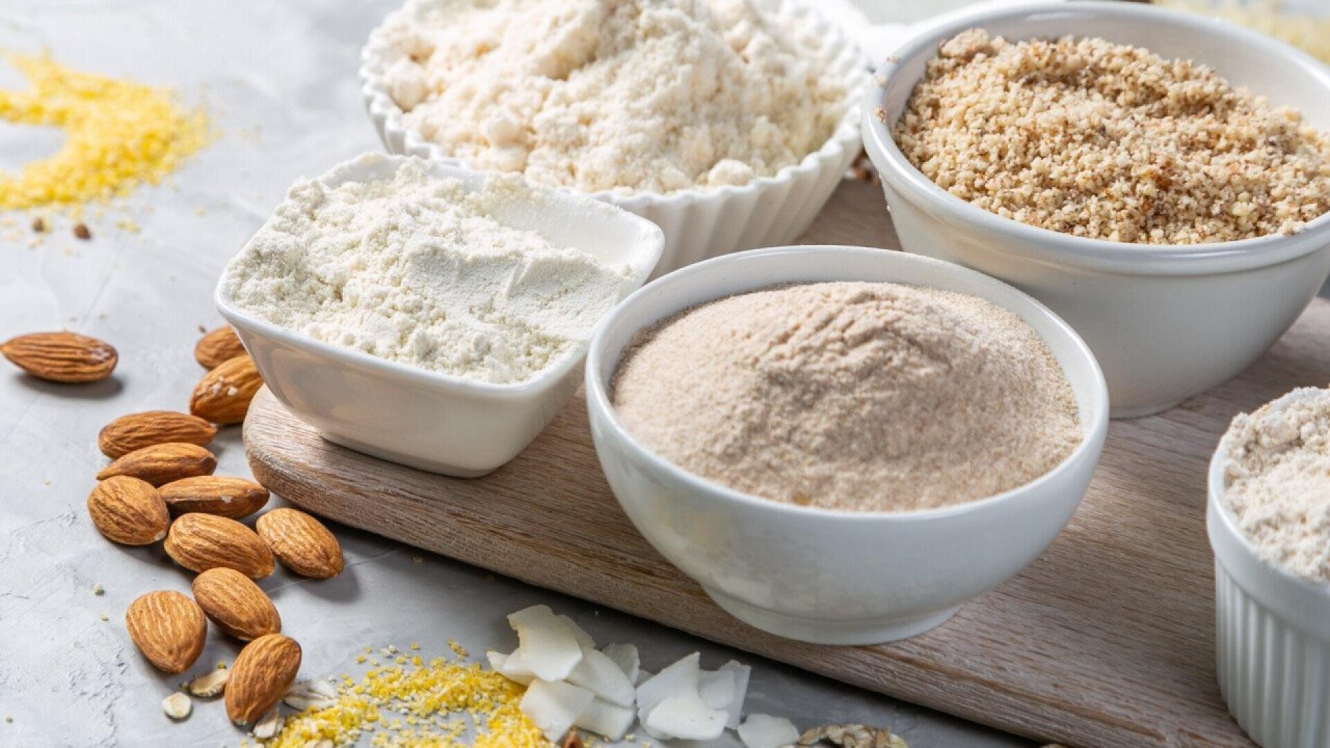 An assortment of gluten-free flours in bowls, including almond flour, coconut flour, and oat flour, arranged on a wooden board with whole almonds and coconut flakes scattered nearby.