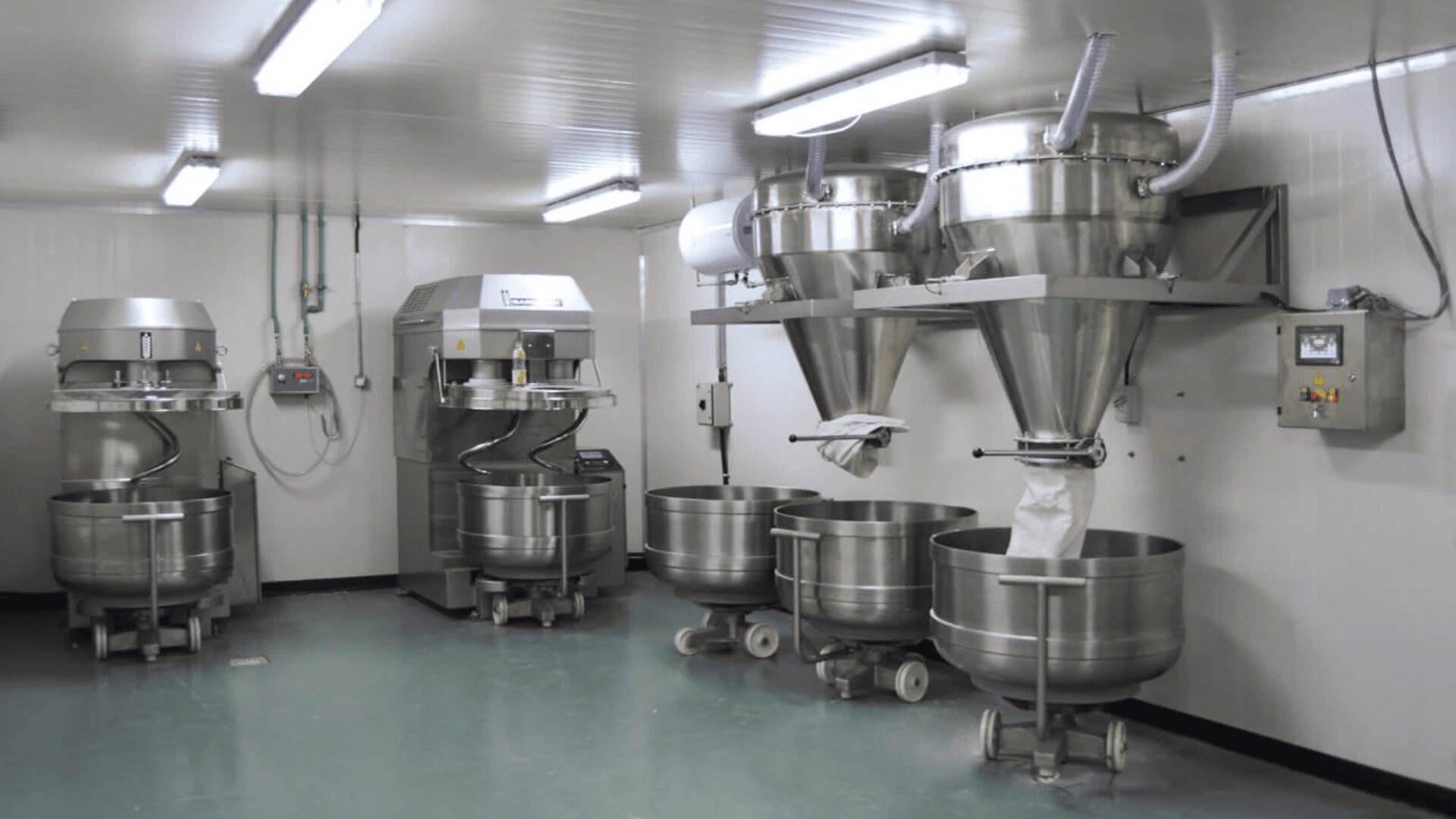 A clean, modern industrial kitchen with large stainless steel mixing machines, metal bowls on wheels, and automated ingredient dispensers attached to the wall, under bright overhead lighting.