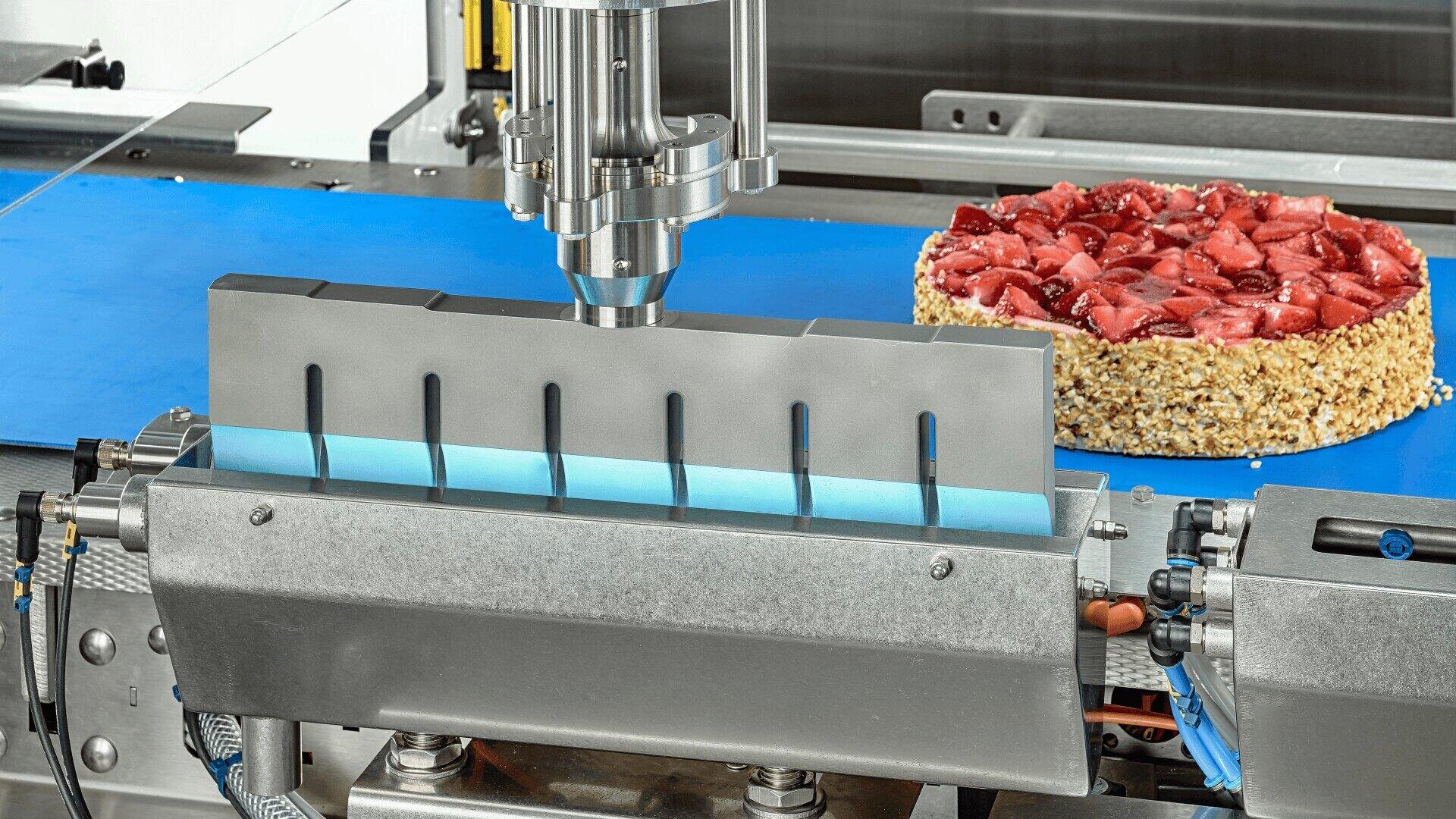 Eine Maschine mit einer Metallklinge schneidet einen runden Erdbeerkuchen mit einem nussfarbenen Rand auf einem blauen Förderband in einer Lebensmittelverarbeitungsanlage.