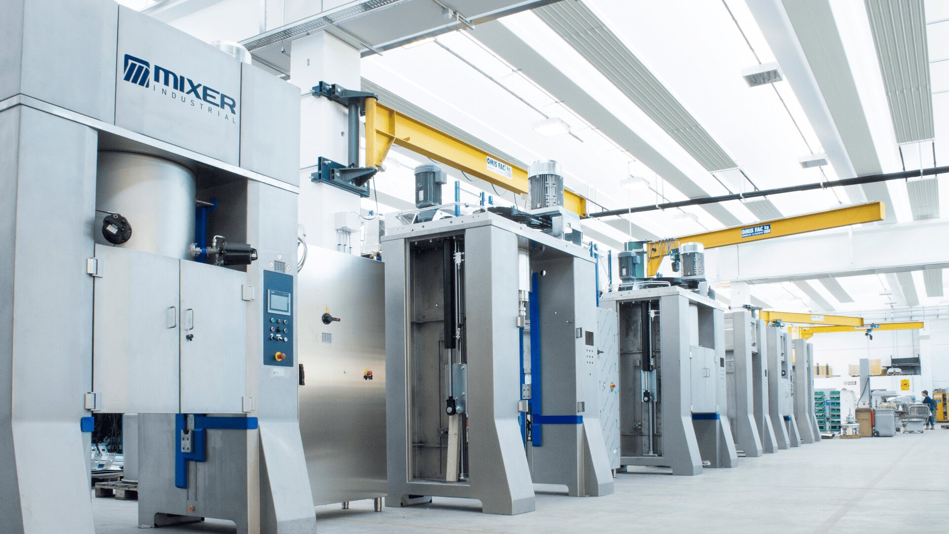 A row of large, stainless steel industrial machines with control panels in a bright, modern factory setting. Overhead cranes and lights are visible above the equipment.