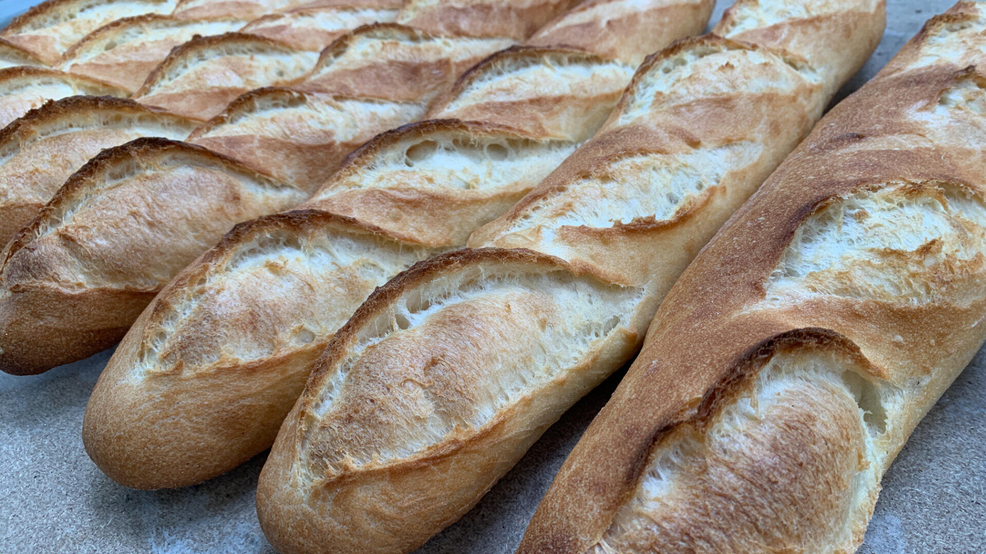 Several freshly baked baguettes with a golden, crispy crust lie in rows on a flat surface. The bread has characteristic diagonal cuts and looks warm and inviting.