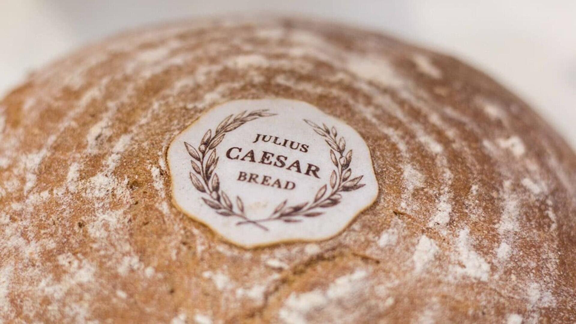 Nahaufnahme eines runden Brotlaibs mit einem Etikett mit der Aufschrift Julius-Cäsar-Brot, umgeben von einem Lorbeerkranz-Muster. Das Brot hat eine leicht bestäubte, rustikale Kruste.
