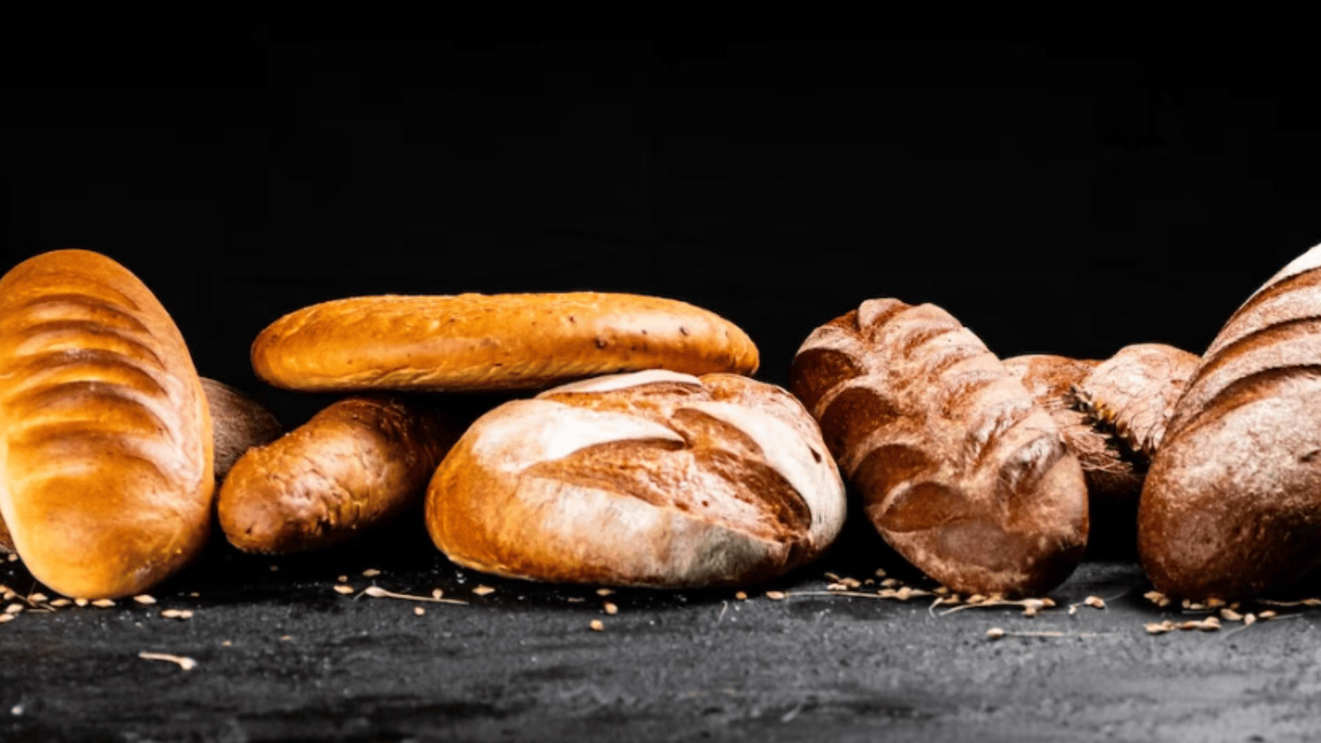 Auf einer schwarzen Fläche vor einem dunklen Hintergrund sind verschiedene Brotlaibe und Baguettes angeordnet, um die herum einige Weizenkörner verstreut sind.