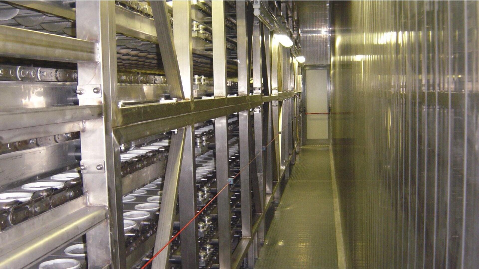 Ein schmaler Gang führt entlang von Metallregalen, die mit Reihen von gestapelten Tabletts oder Behältern gefüllt sind, in einer Industrie- oder Lagerhalle mit heller Beleuchtung.