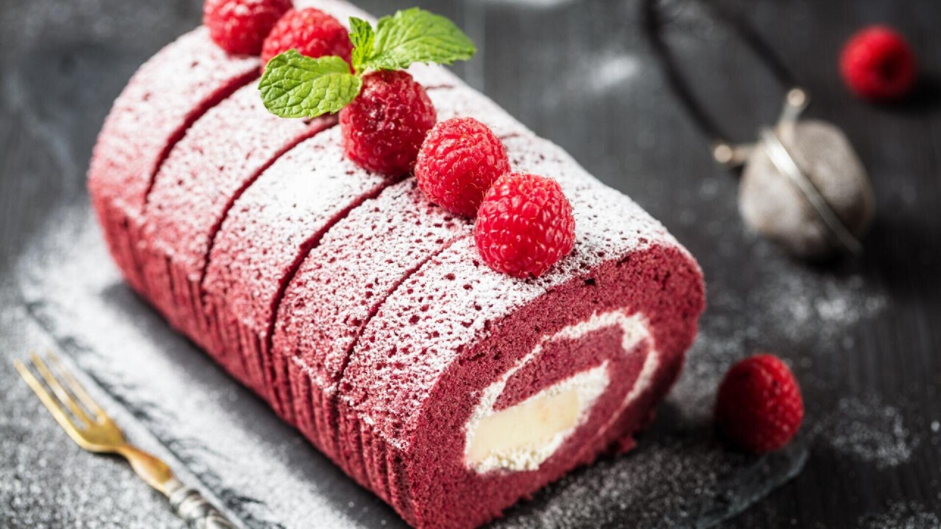 A sliced red velvet cake roll dusted with powdered sugar, filled with cream, and topped with fresh raspberries and a sprig of mint, displayed on a dark surface with a fork nearby.