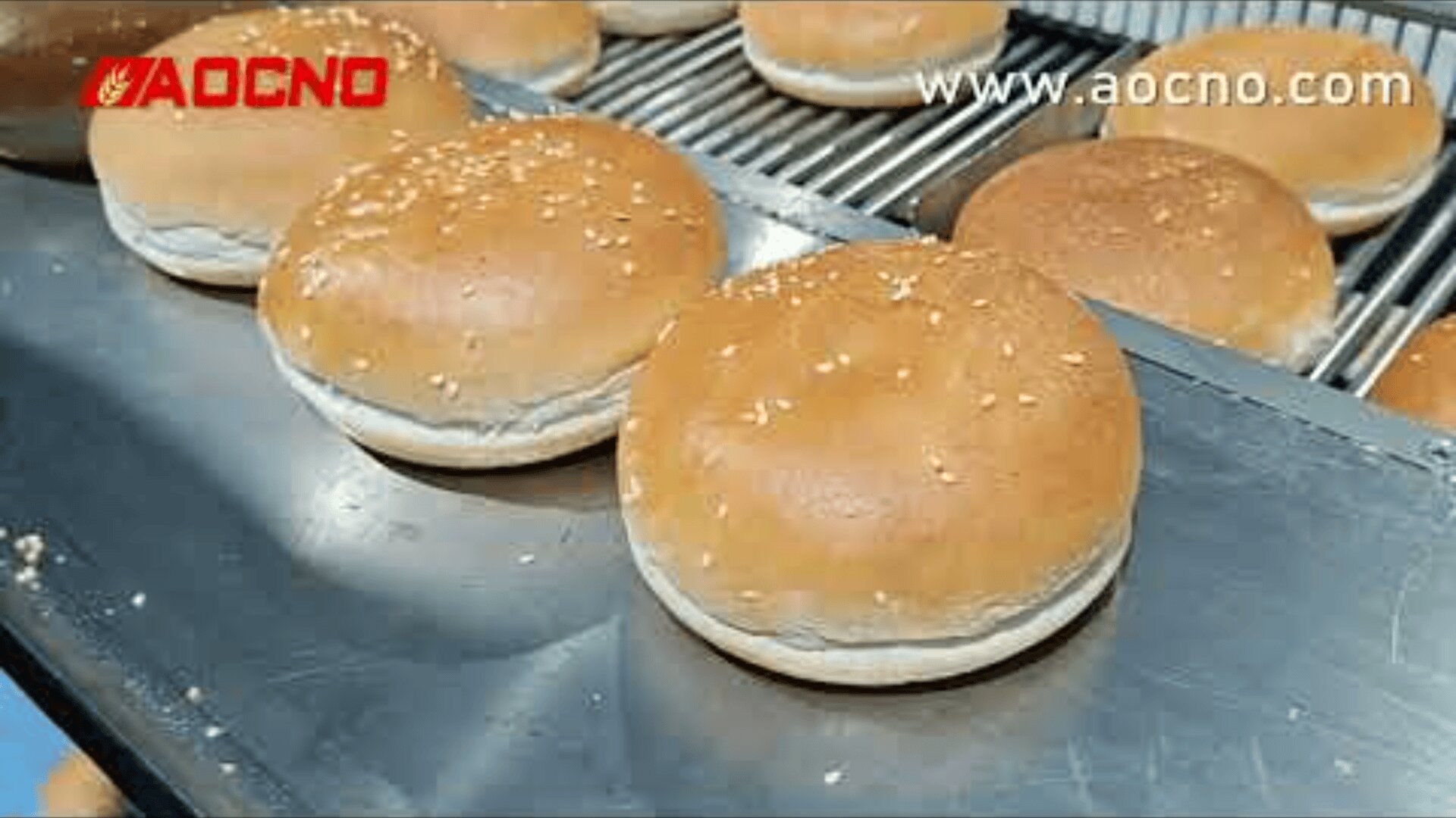 Several freshly baked hamburger buns with sesame seeds are lined up on a metal conveyor belt in a bakery. The logo AOCNO and the website www.aocno.com appear in the image.