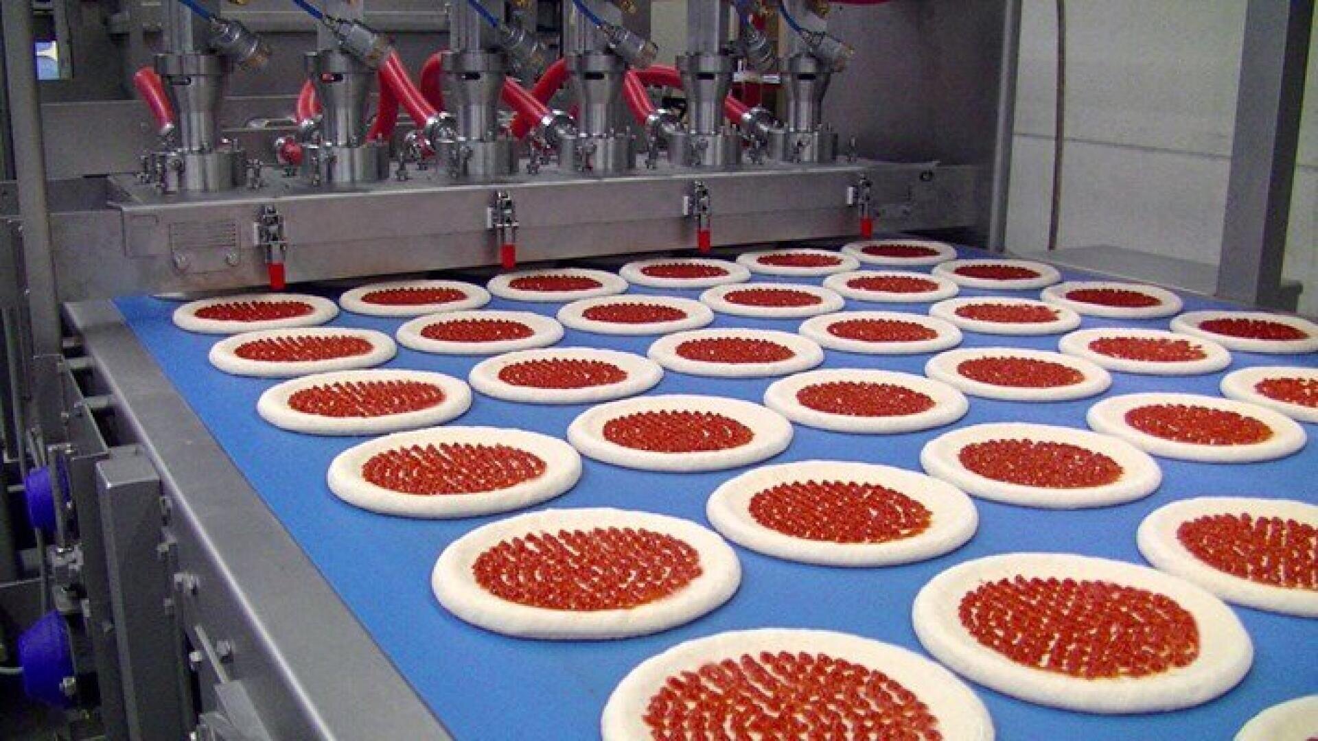 Rows of raw pizzas with sauce are arranged on a blue conveyor belt and run through an automatic machine that spreads the sauce in precise circles on each dough base in a food production line.
