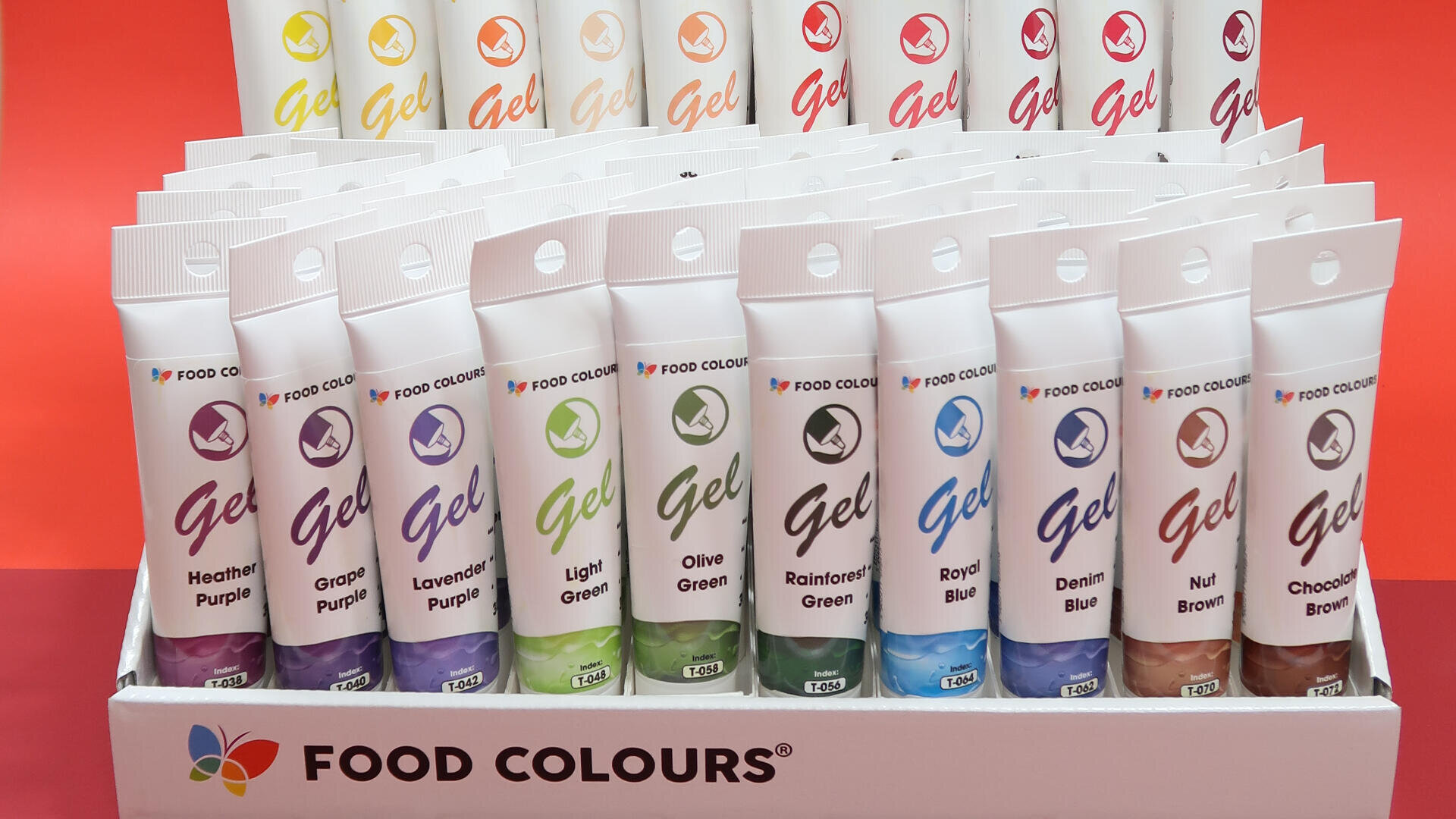 A display box of FOOD COLOURS branded gel food coloring tubes in various shades, including purple, green, blue, and brown, arranged upright against a red background.