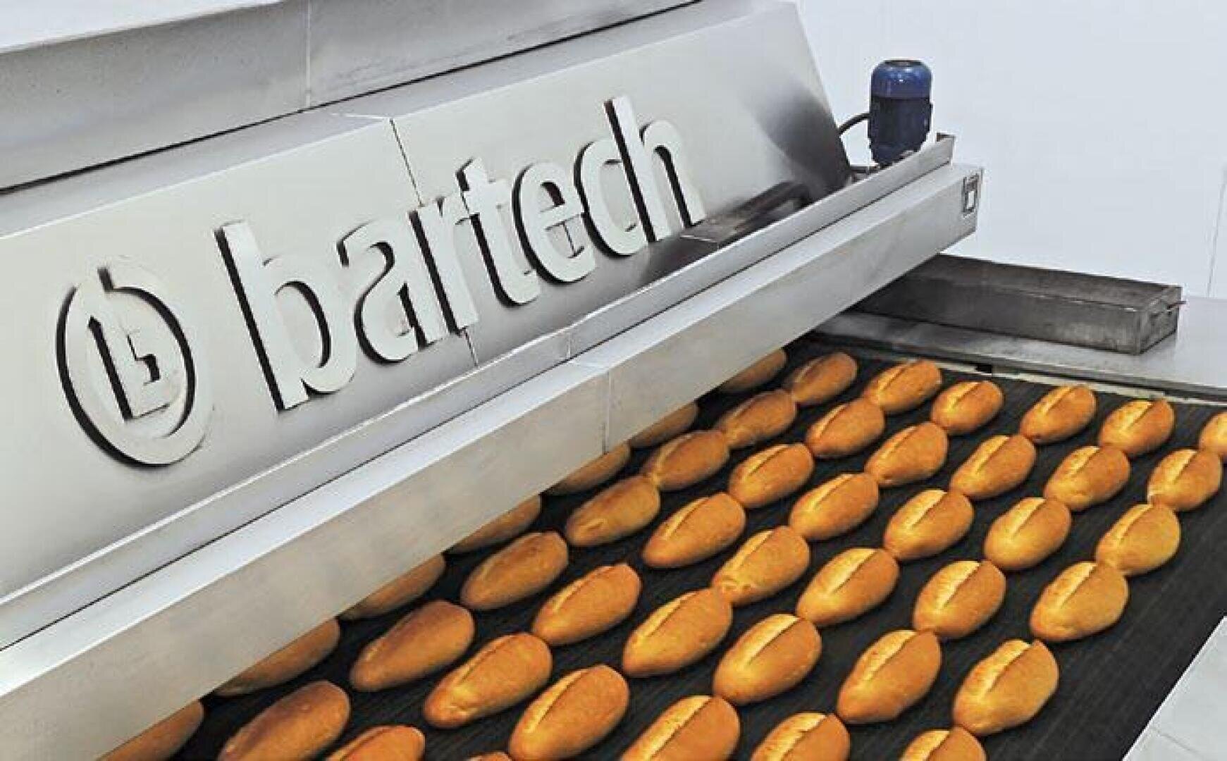 In einer modernen Bäckerei laufen goldbraune Brötchen über ein Förderband unter einer Edelstahlmaschine mit der Aufschrift "bartech".