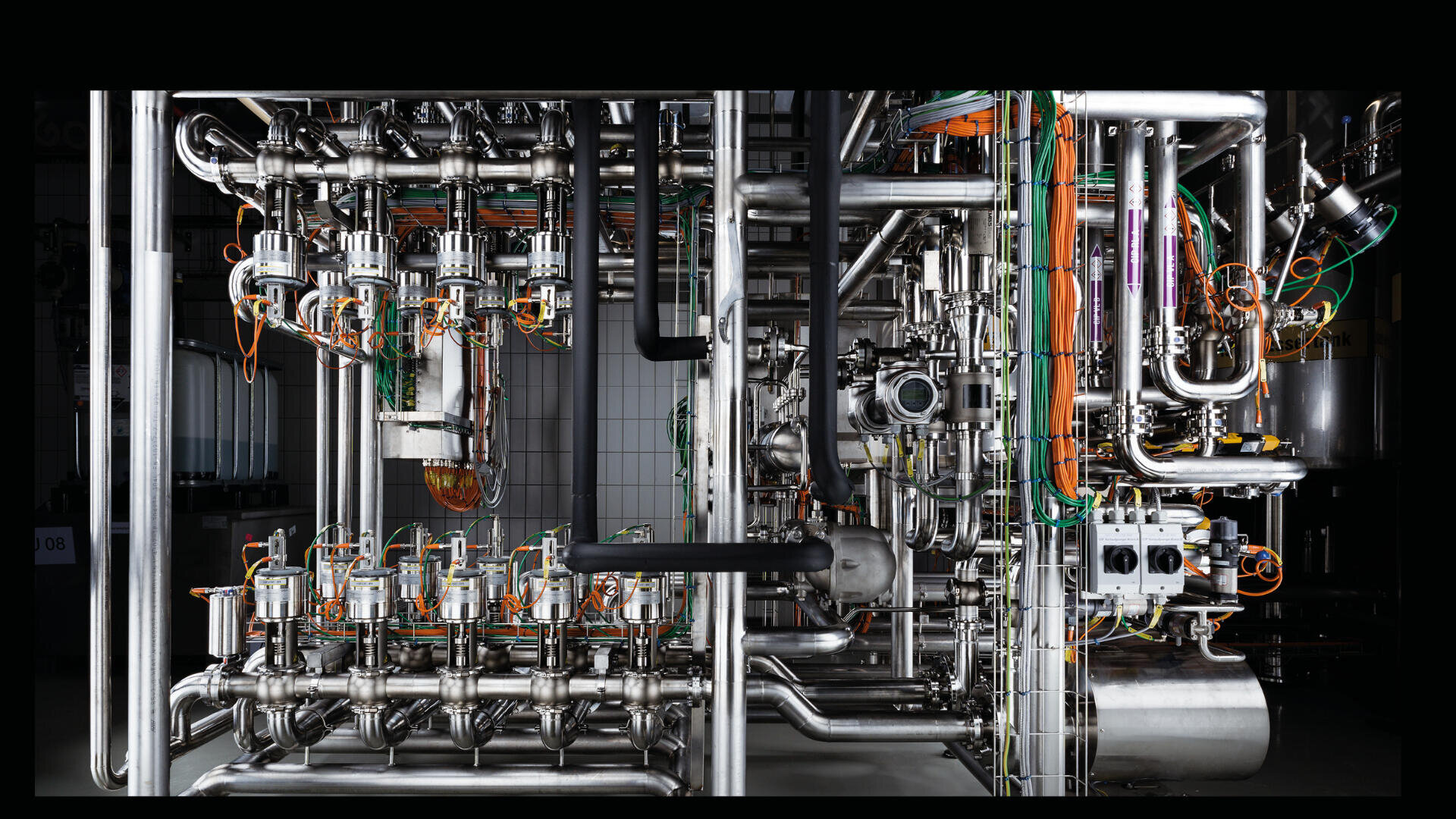 Ein komplexes Netzwerk aus industriellen Edelstahlrohren, Ventilen und Kabeln mit orangefarbenen und grünen Drähten, Maschinen und Schalttafeln in einer sauberen, gut beleuchteten Anlage.