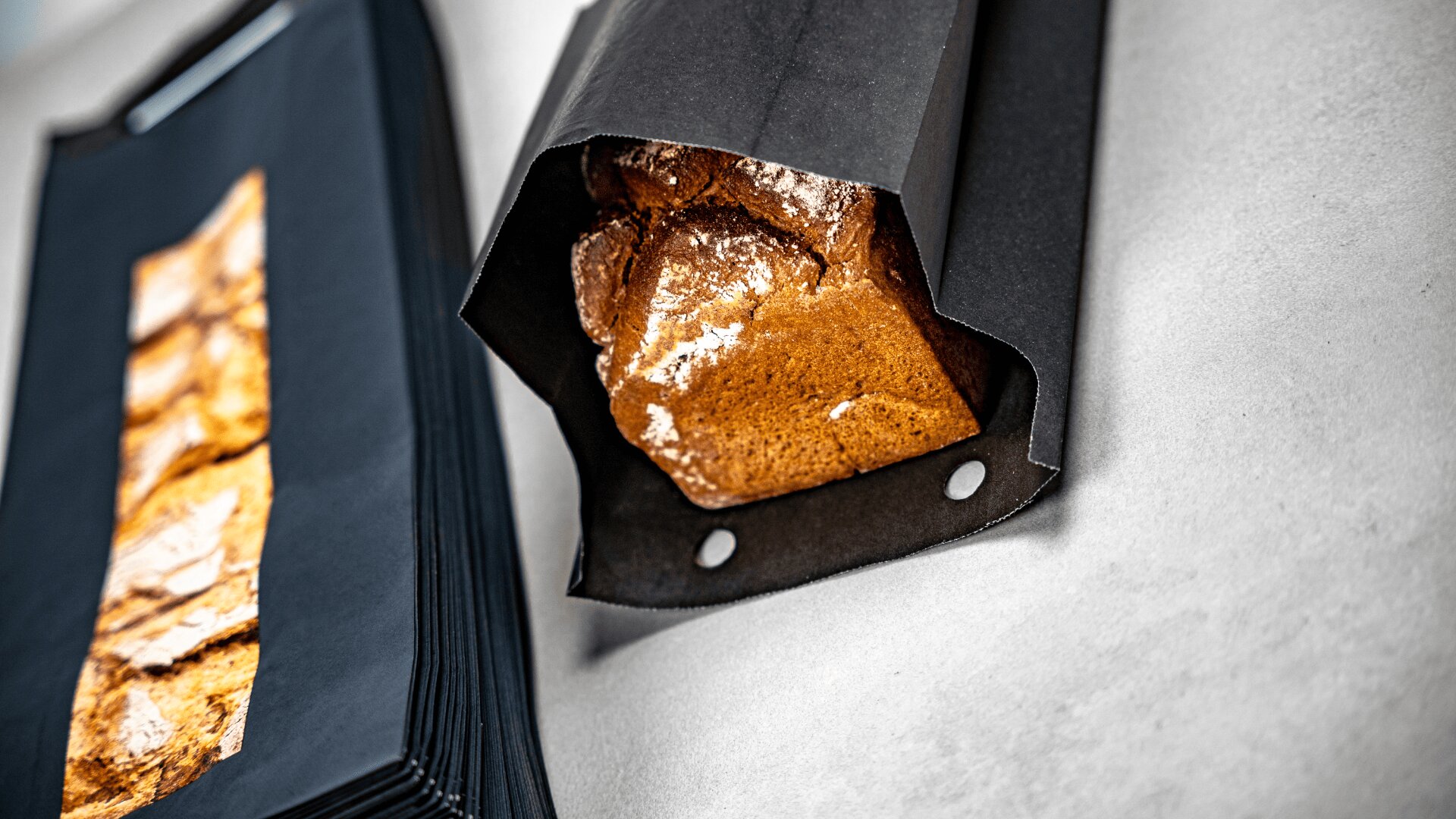 Ein teilweise in schwarzes Papier eingewickelter Brotlaib liegt auf einer hellen Fläche, daneben liegt ein Stapel schwarzer Papierblätter.
