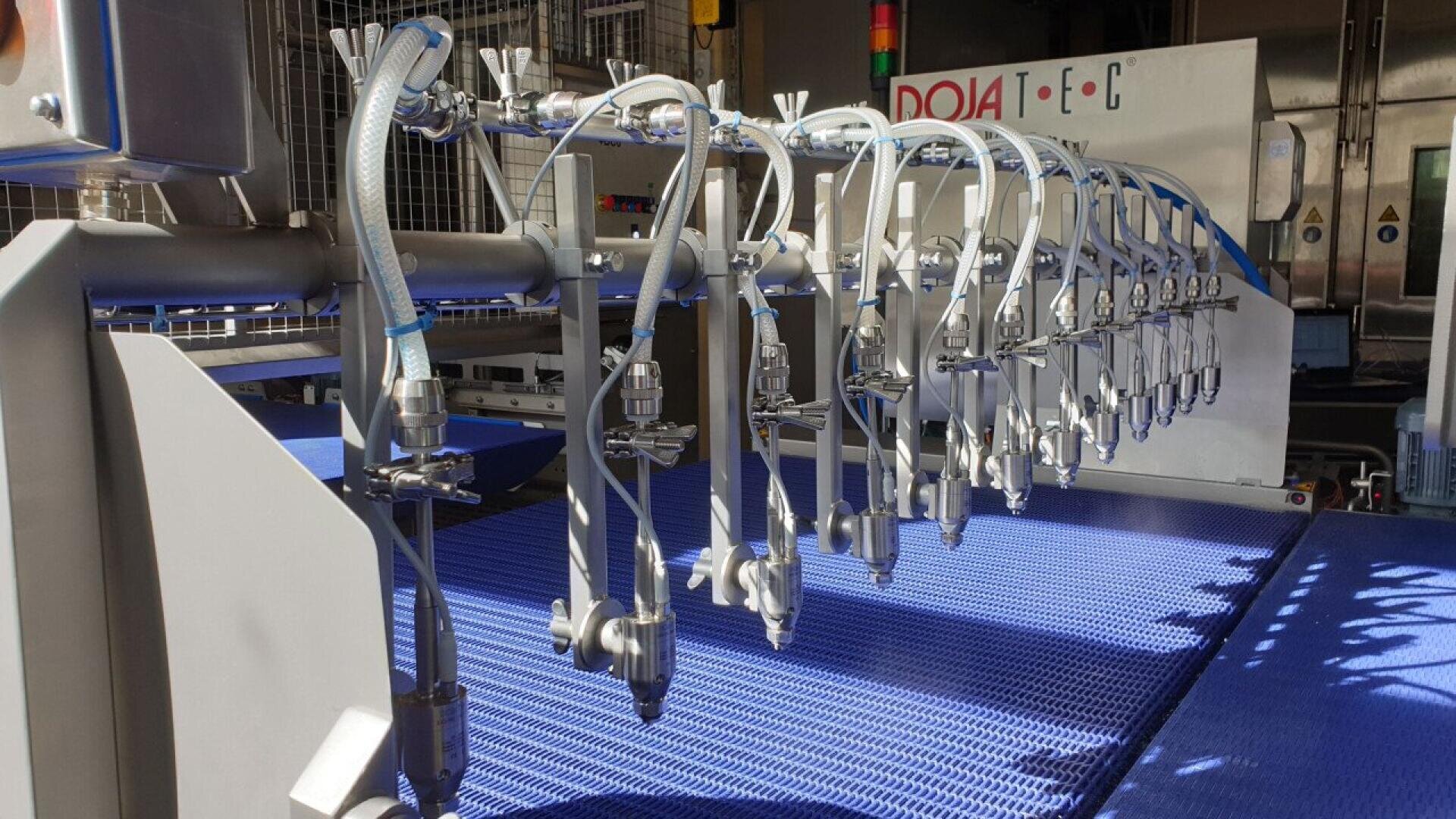 Rows of metal nozzles attached to blue hoses are lined up above a blue conveyor belt in an industrial food processing or manufacturing machine.