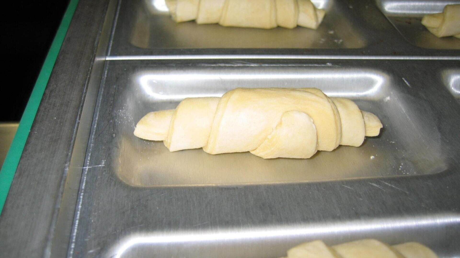 Ungebackener, ausgerollter Hörnchenteig auf einem Metallblech, bereit zum Backen. Der Teig hat eine helle Farbe und liegt in einem der rechteckigen Bereiche des Blechs.