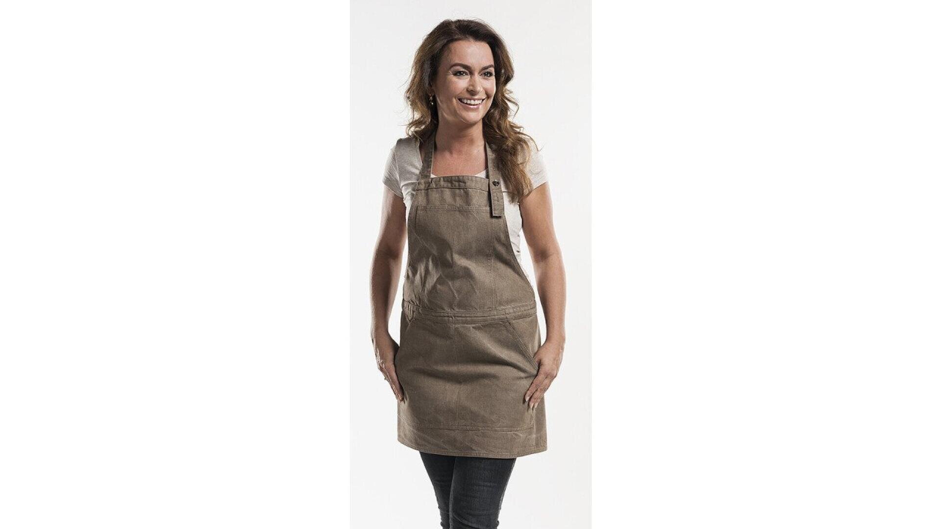 A woman with brown hair, wearing a white shirt and a light brown apron, stands smiling with her hands resting on the sides of her apron against a plain white background.