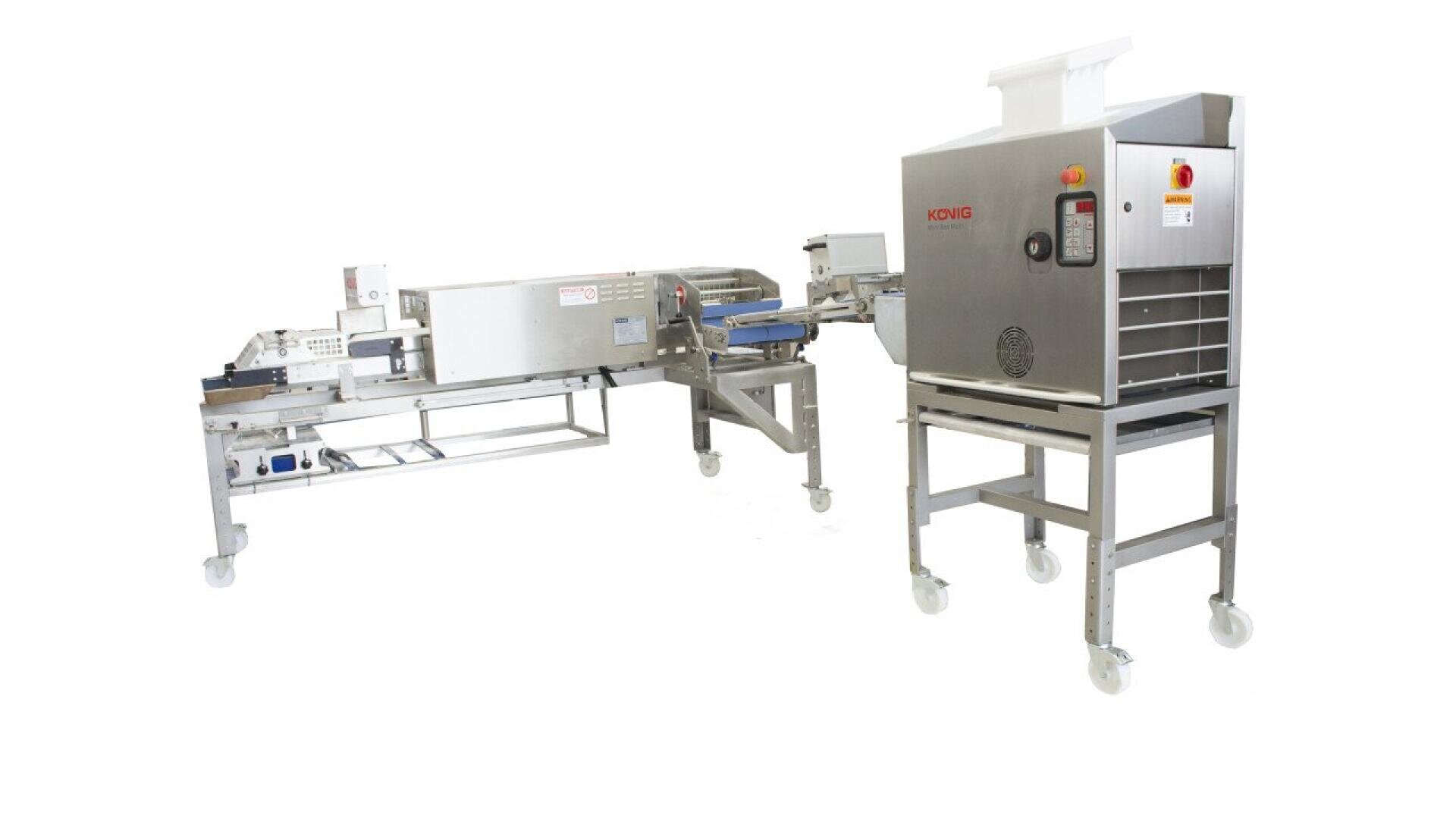 A large industrial dough processing machine made of stainless steel with conveyor belts, control panels, and caster wheels for mobility, used in commercial baking or food production.