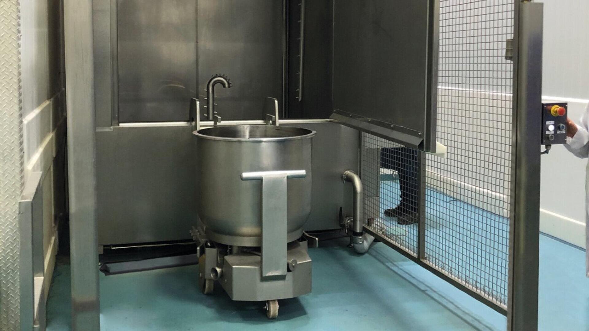 A large stainless steel industrial mixing bowl on wheels is positioned inside a metal enclosure with a mesh safety door. A person’s hand is pressing a red emergency stop button on a control panel outside the enclosure.