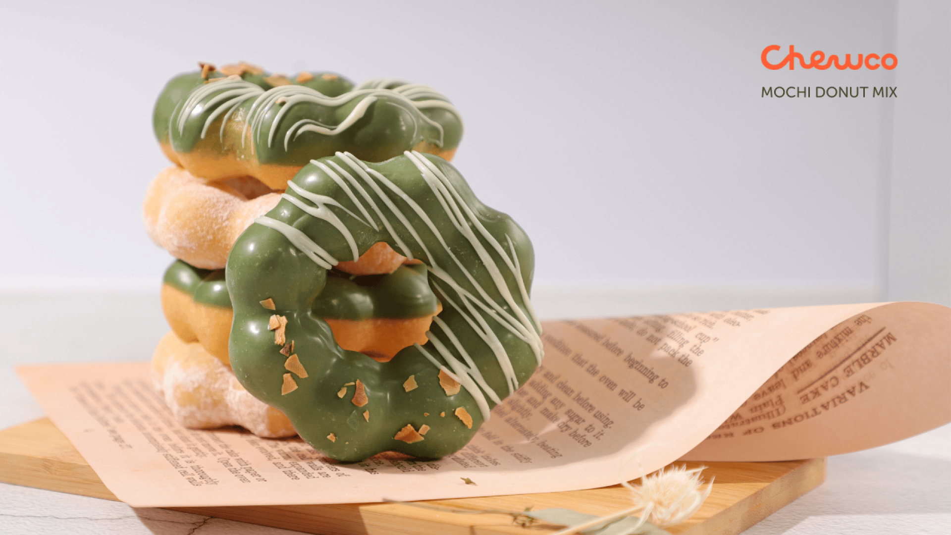 Three mochi donuts with green matcha glaze and white drizzle are stacked on pages of a book. The donuts are labeled “Chewco MOCHI DONUT MIX” in the top right corner. A small dried flower sits nearby.