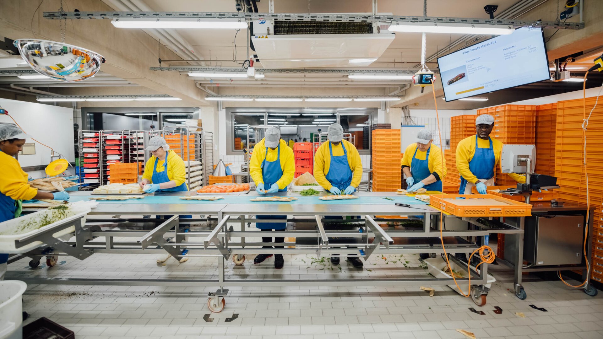 Sechs Mitarbeiter in gelben Hemden, blauen Schürzen und Haarnetzen bereiten an einem langen Tisch in einer hell erleuchteten Industrieküche mit orangefarbenen Tabletts auf Regalen und einem Monitor über dem Kopf das Essen zu.
