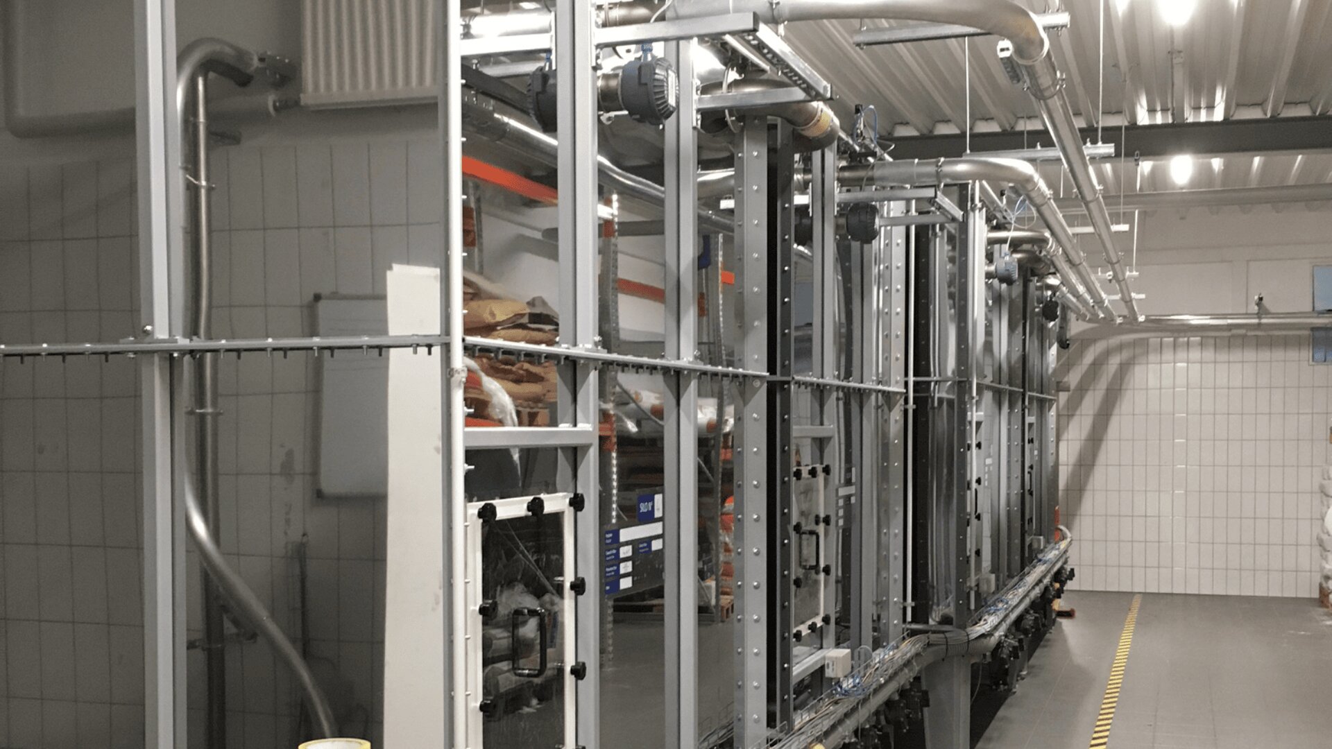 Industrielle Verarbeitungsanlagen in einem gefliesten Raum mit Metallrohren, Gestellen und Maschinen entlang eines Fördersystems; die gelbe gestrichelte Linie markiert den Weg auf dem Boden.