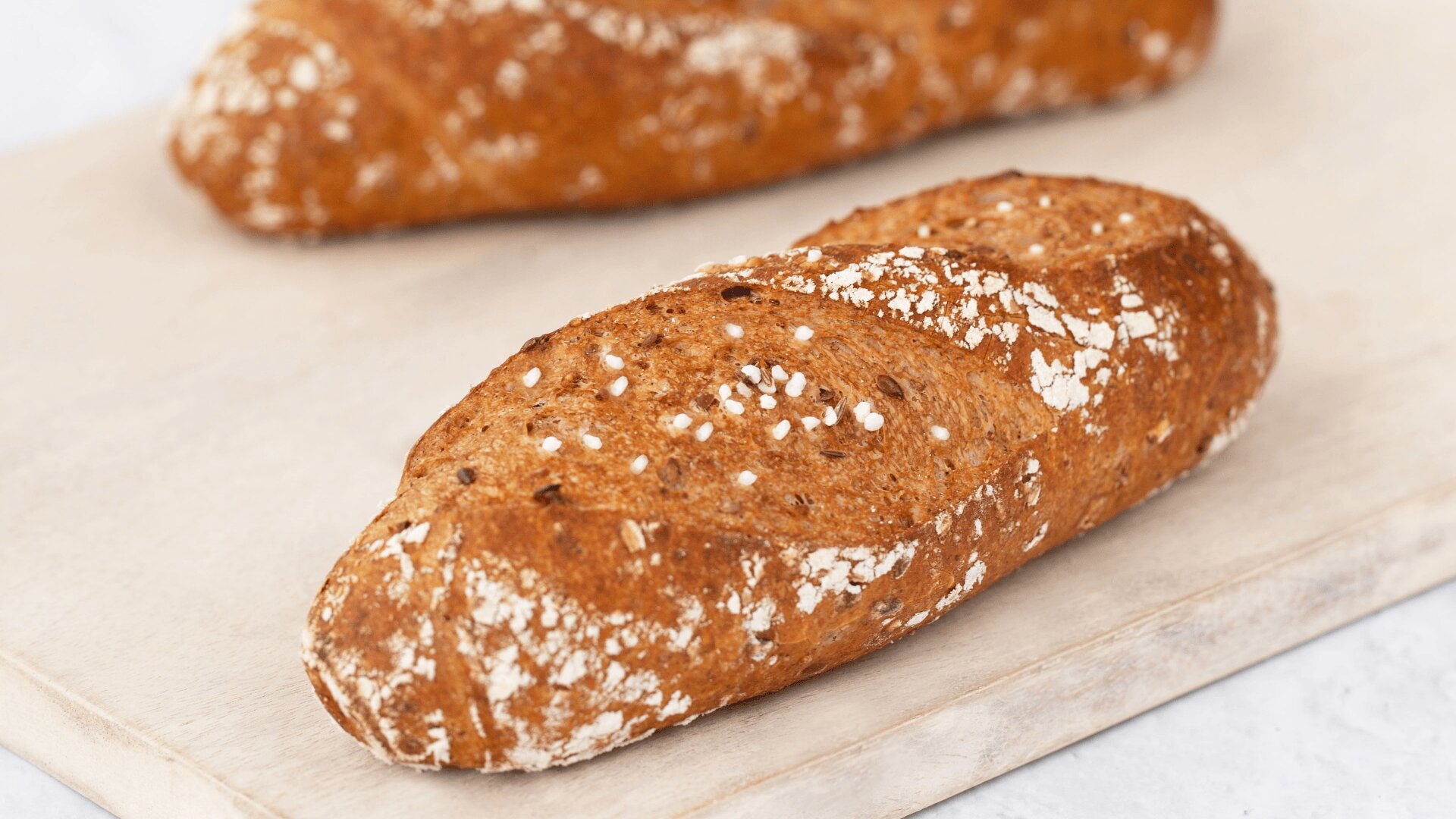 Nahaufnahme eines rustikalen, ovalen Vollkornbrötchens mit knuspriger, goldbrauner Kruste und mit Hafer bestreut, das auf einer hellen Holzunterlage ruht. Ein anderes ähnliches Brötchen ist im Hintergrund verschwommen.