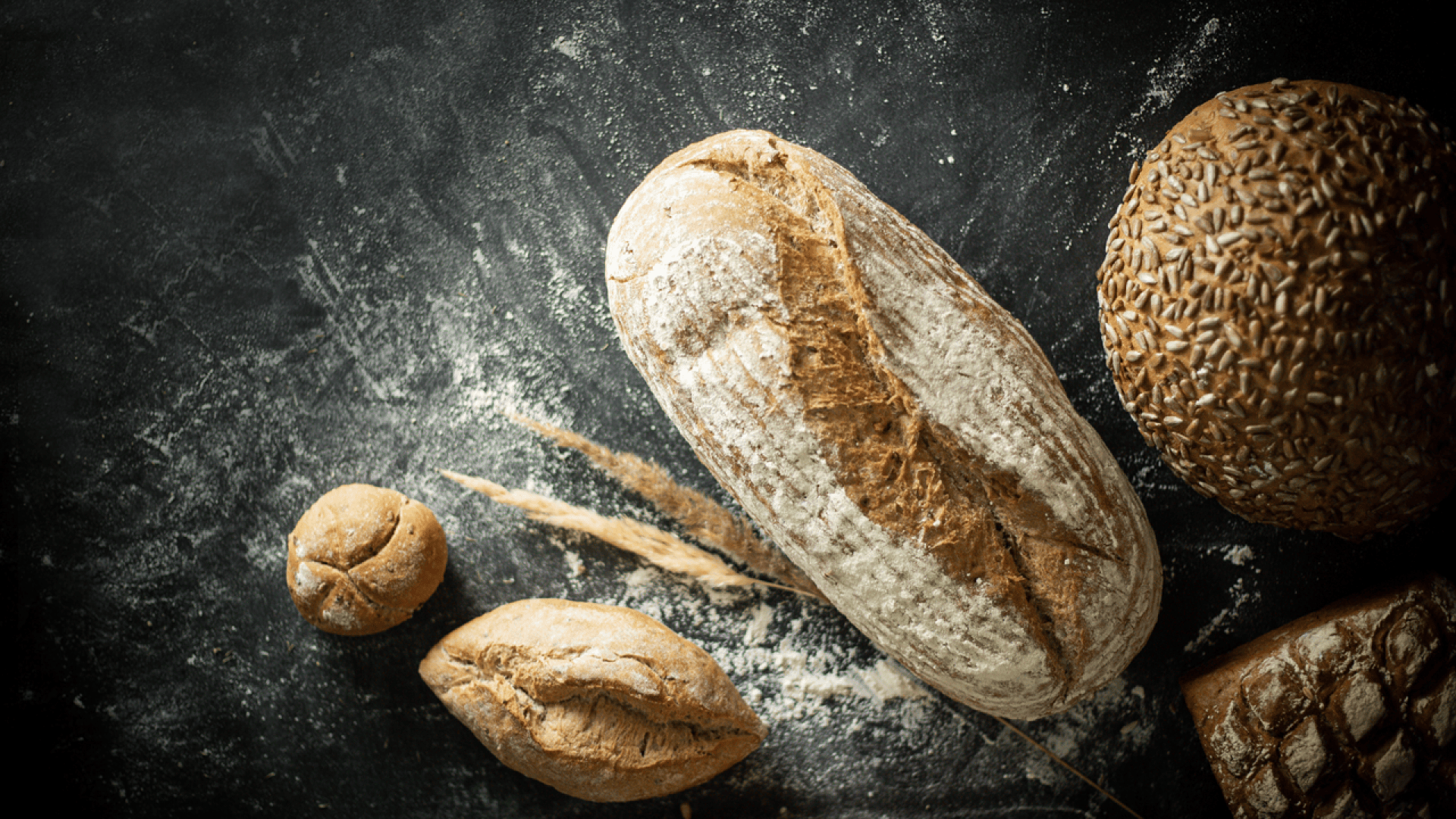 Assorted artisan breads, including a round loaf with seeds and a long rustic loaf, are arranged on a dark, floured surface.