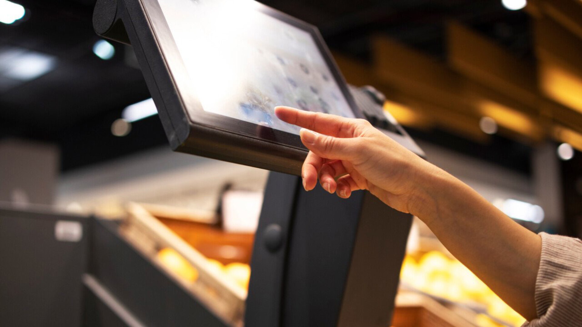 A person’s hand reaches out to touch a touchscreen kiosk in a store, with blurred shelves of products visible in the background.