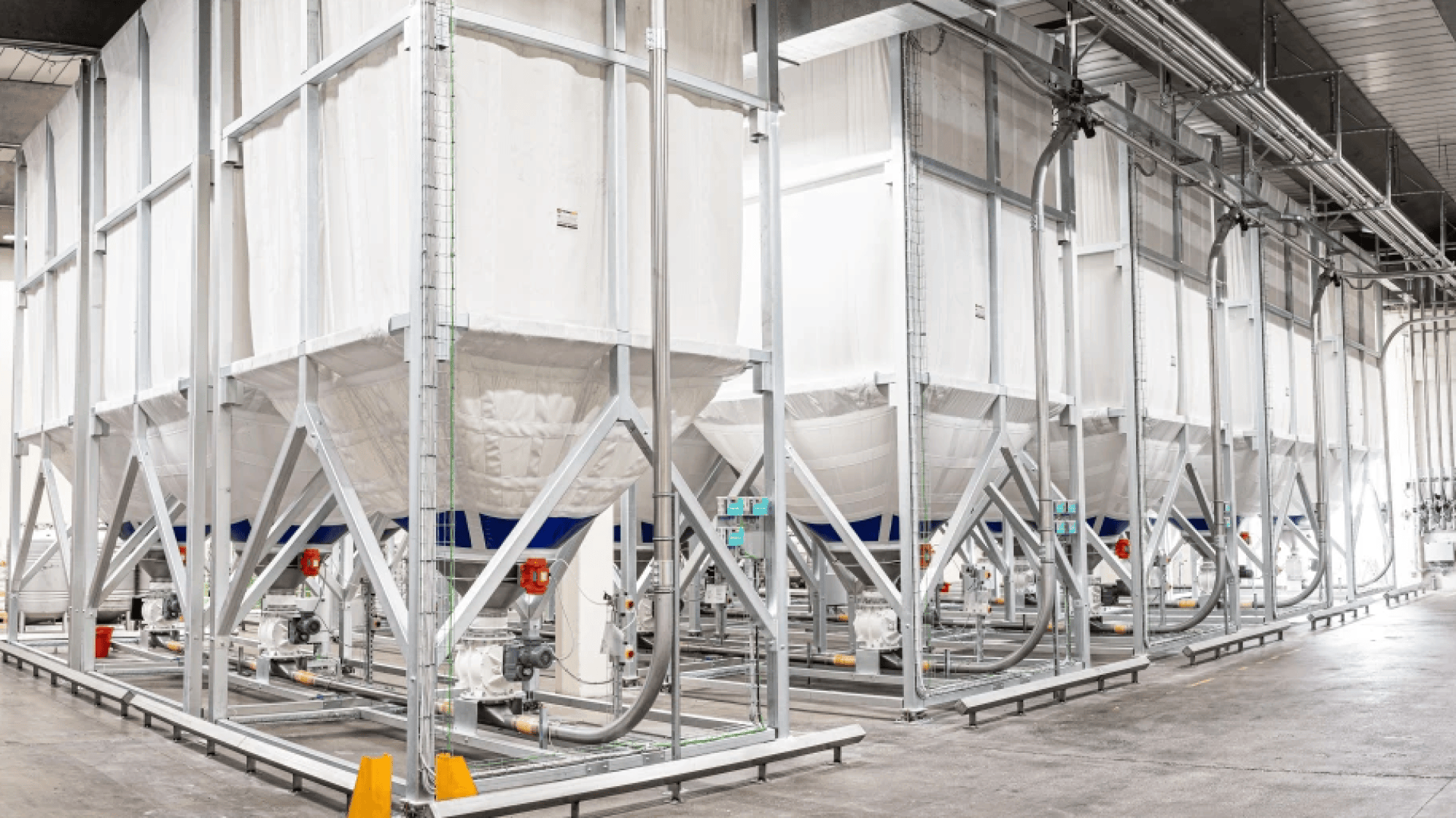 Large white industrial storage silos or bins with metal frames and pipes are lined up inside a bright, clean factory or processing facility. The floor is concrete and there are various valves and machinery connected to the bins.