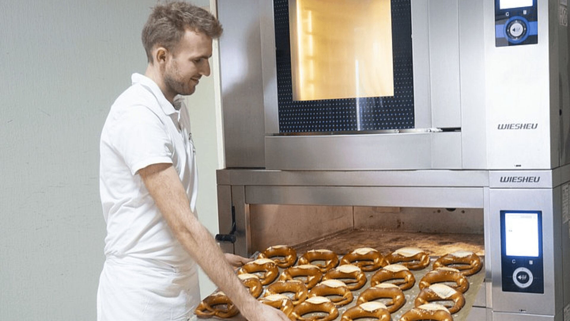 Ein Bäcker in weißer Uniform entnimmt ein Tablett mit frisch gebackenen Brezeln aus einem großen Industrieofen in einer Bäckerei.