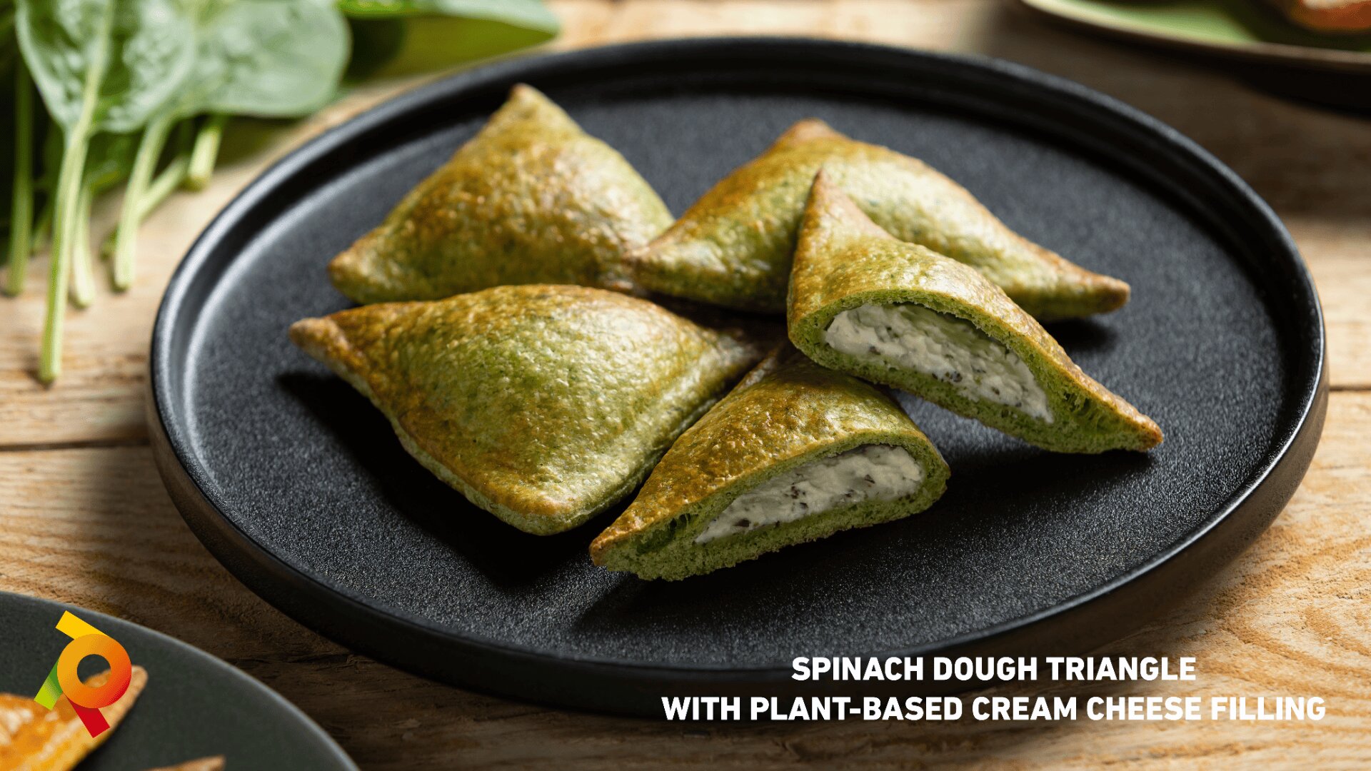 Four spinach dough triangles with a creamy white plant-based filling are arranged on a black plate, with fresh spinach leaves in the background. Text reads: Spinach Dough Triangle with Plant-Based Cream Cheese Filling.