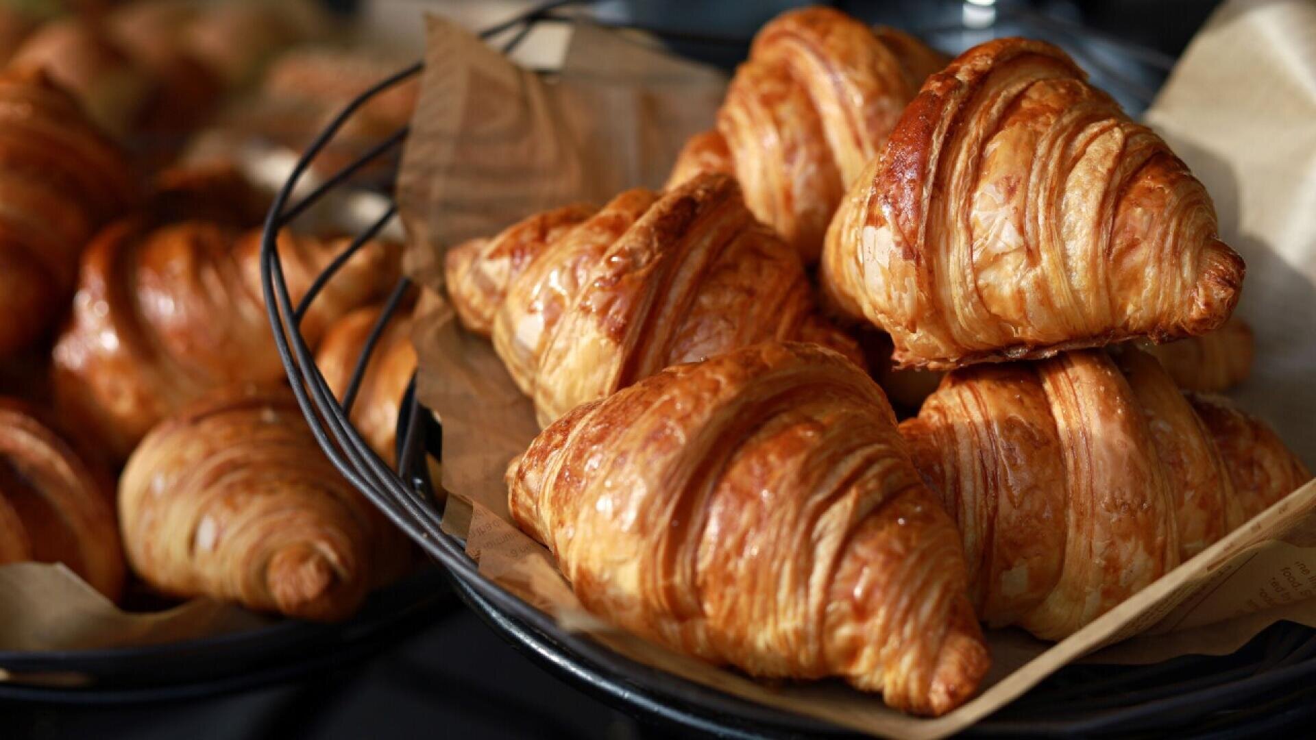 A wire basket lined with brown parchment paper is filled with golden, flaky croissants. More croissants are visible in the background, slightly out of focus.