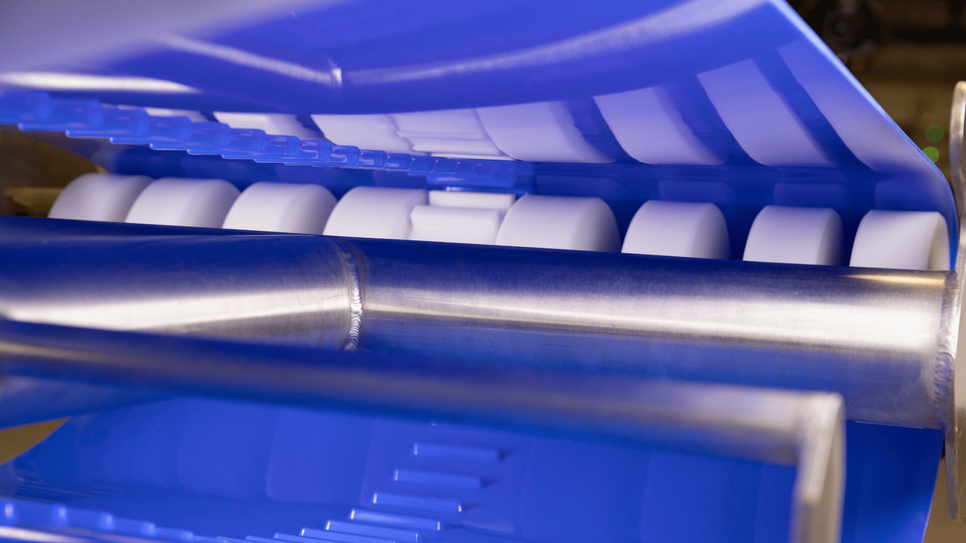 Close-up of a conveyor system with cylindrical rollers and a blue-tinted light, likely used in a manufacturing or food processing facility.