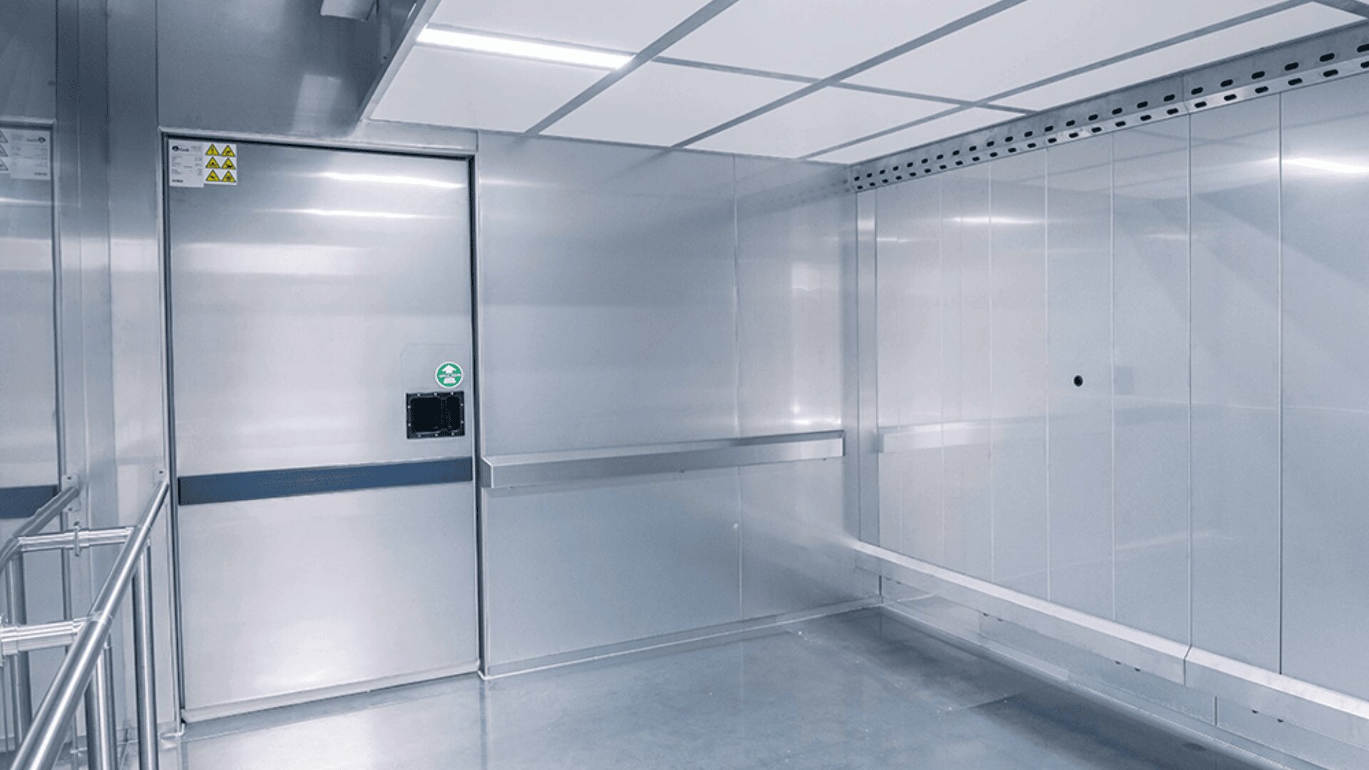 Ein sauberer, leerer Raum aus rostfreiem Stahl mit glatten Wänden, heller Deckenbeleuchtung und einer geschlossenen Schiebetür, wie man sie üblicherweise in Laboratorien oder sterilen Umgebungen findet.